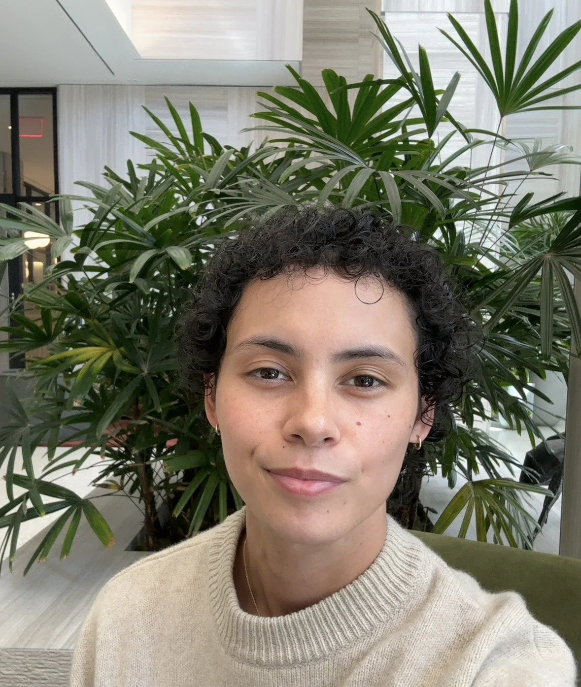 Person wearing a cream sweater indoors, seated in front of lush green plants.