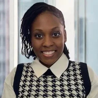 A smiling person wearing a white collard blouse under a black-and-white houndstooth sweater vest, with short styled hair and a softly blurred window background.