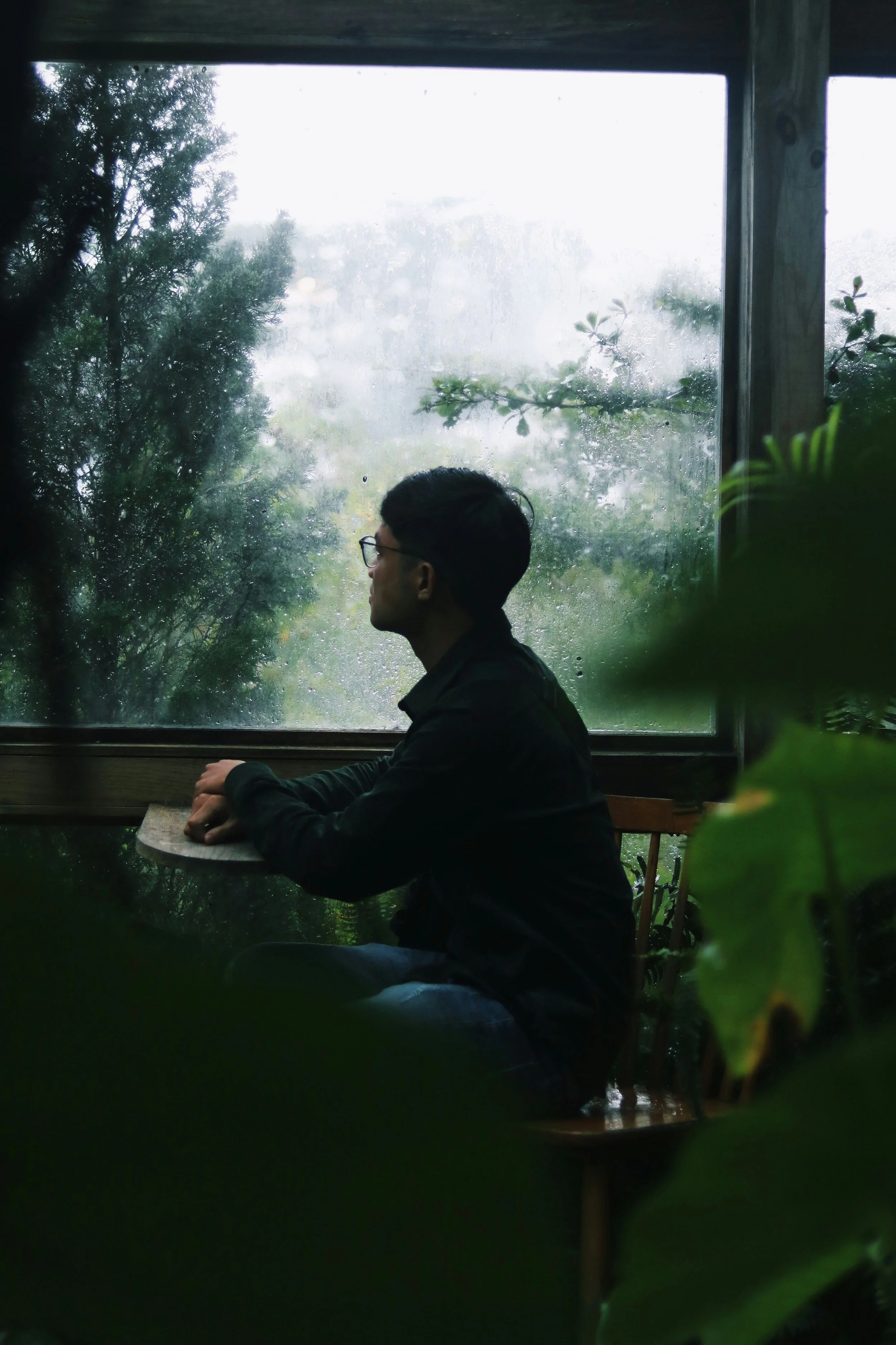 A person sits quietly at a small table indoors, gazing out a large rain-speckled window toward trees and greenery outside, creating a reflective and subdued mood framed by soft natural light.