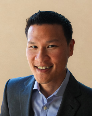 Headshot of Ricky Weng, LMSW smiling broadly at the camera, with short dark hair styled up, wearing a charcoal suit jacket over a light blue open-collar shirt against a warm background.