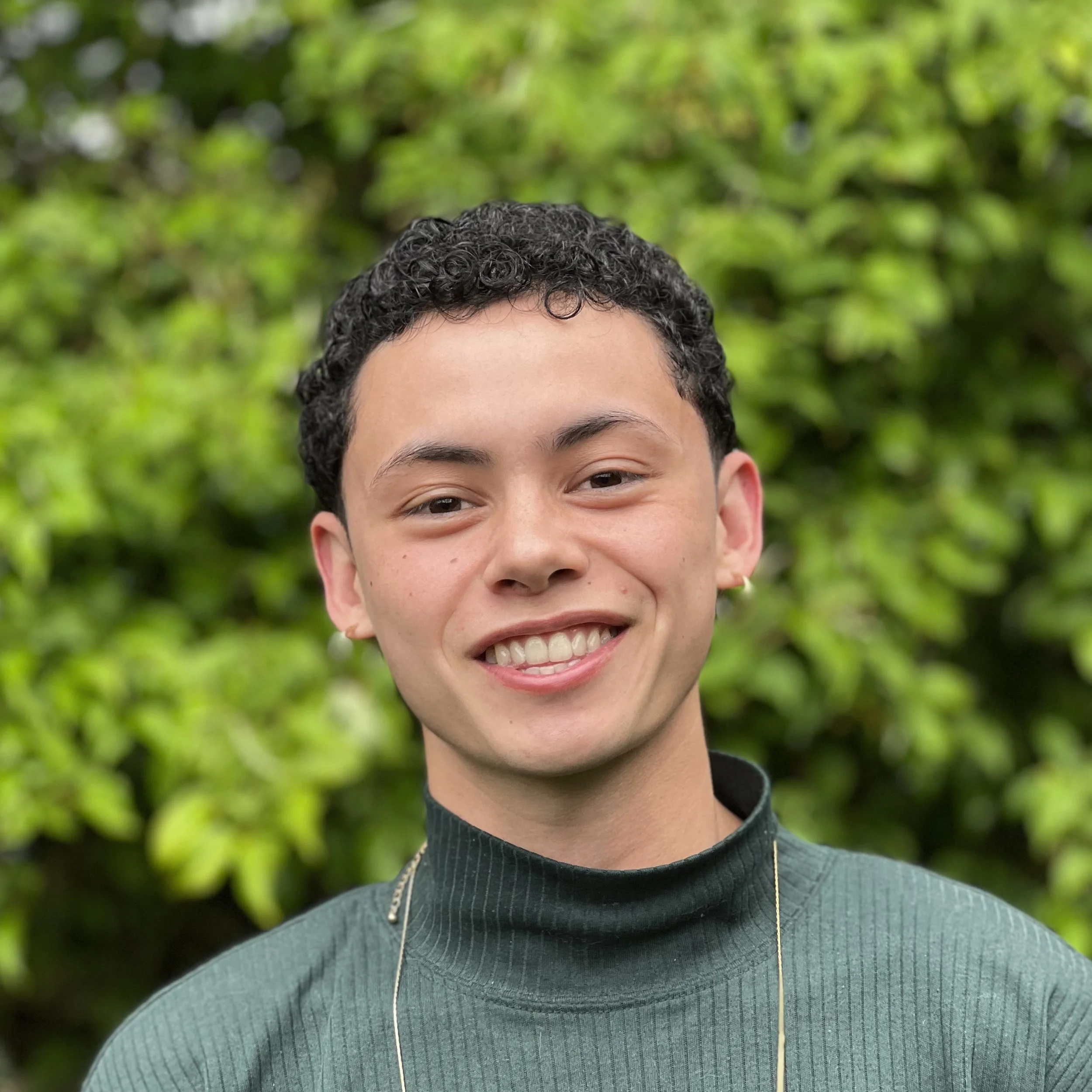 Smiling therapist wearing a dark green turtleneck, photographed outdoors in front of leafy greenery.