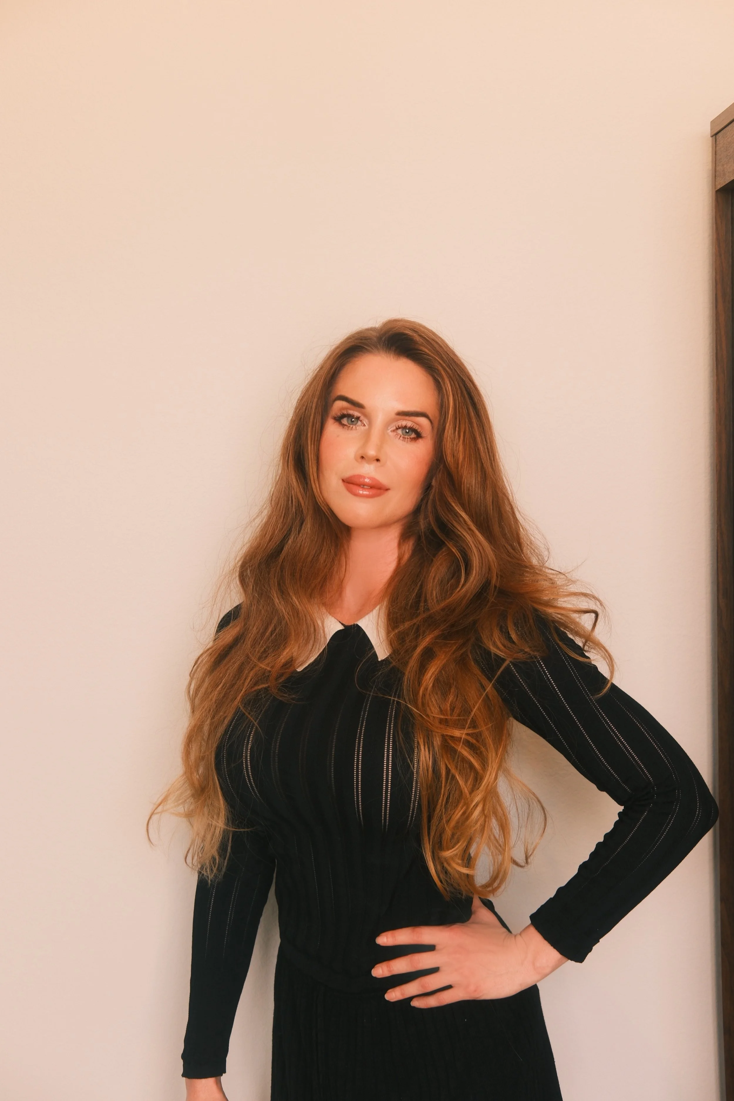 A woman with long, wavy auburn hair stands against a plain cream-colored wall, wearing a fitted black dress with subtle vertical detailing and a white collar. She faces the camera with a soft, confident expression, one hand resting on her hip.