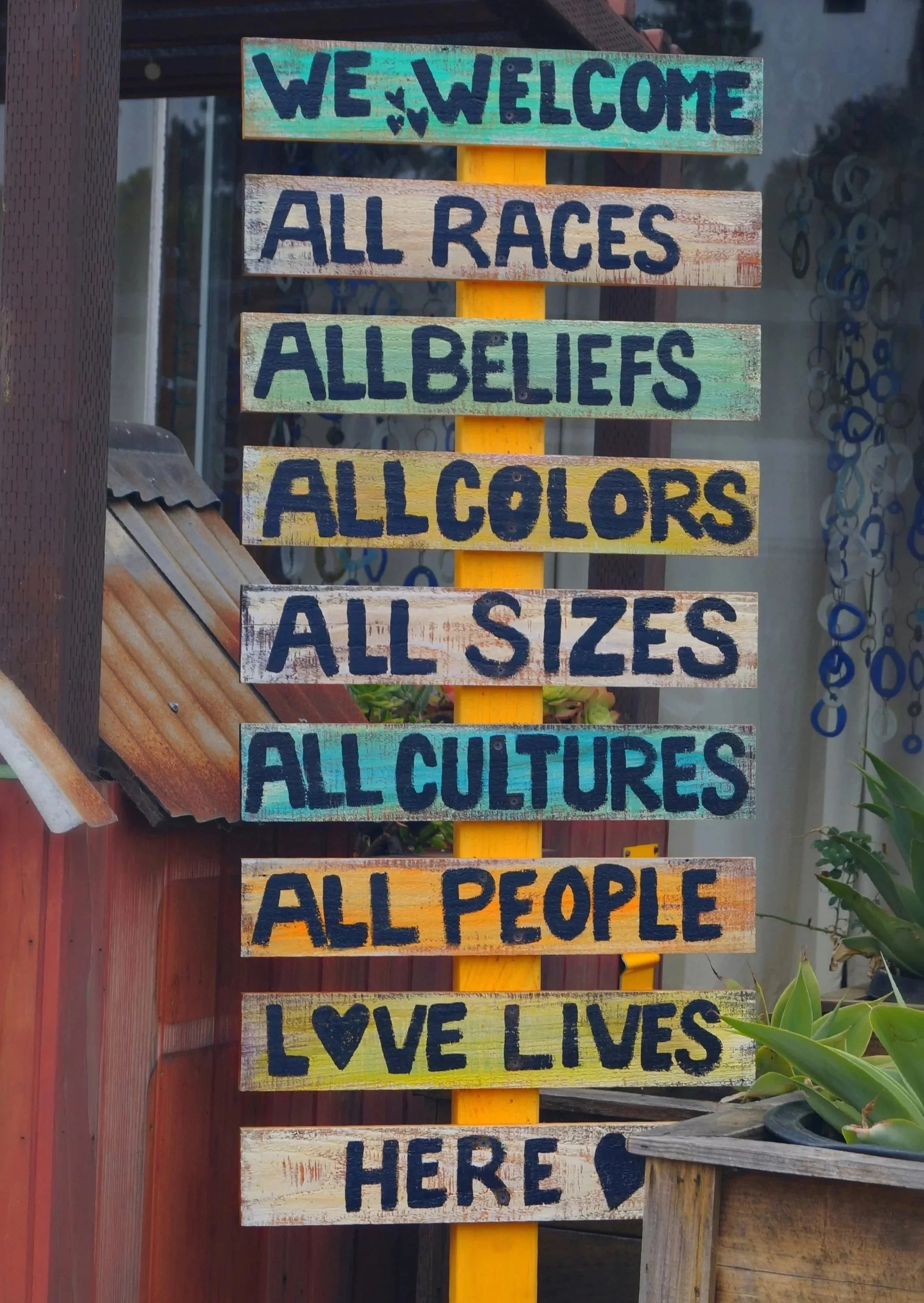 Colorful wooden signpost outside a building reads, “We welcome all races, all beliefs, all colors, all sizes, all cultures, all people—love lives here.
