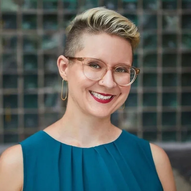Smiling person with a short haircut with blonde tips and brown roots wearing glasses and red lipstick with gold dangle earrings. The person is wearing a teal shirt and is standing in front of a checkered back drop.