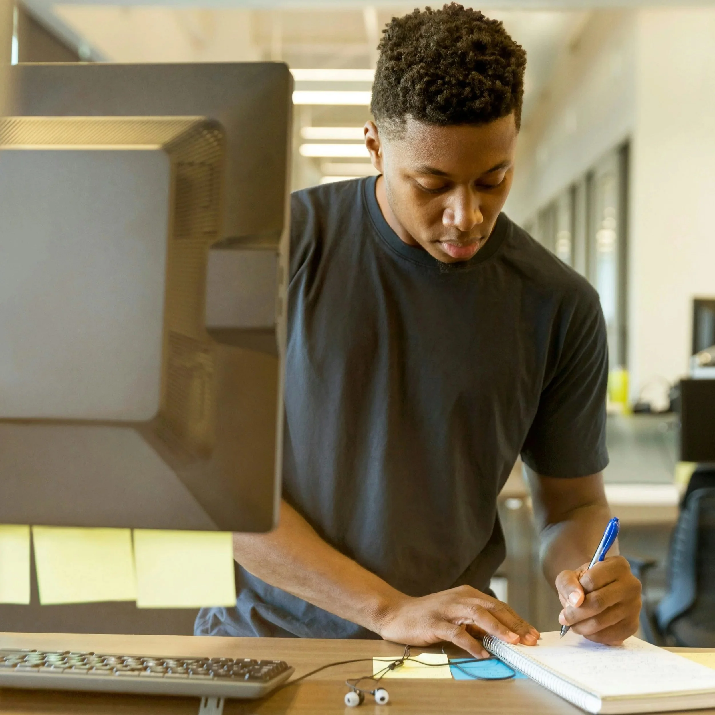 Person standing at an office desk, writing in a notebook beside a computer monitor.