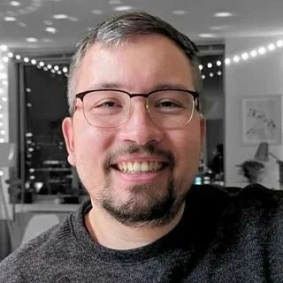 A smiling man with glasses and gray hair, indoors with string lights and a framed picture in the background.