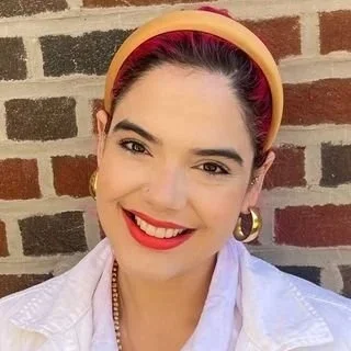 Young woman with red lipstick, hoop earrings, and a yellow headband smiling in front of a brick wall.
