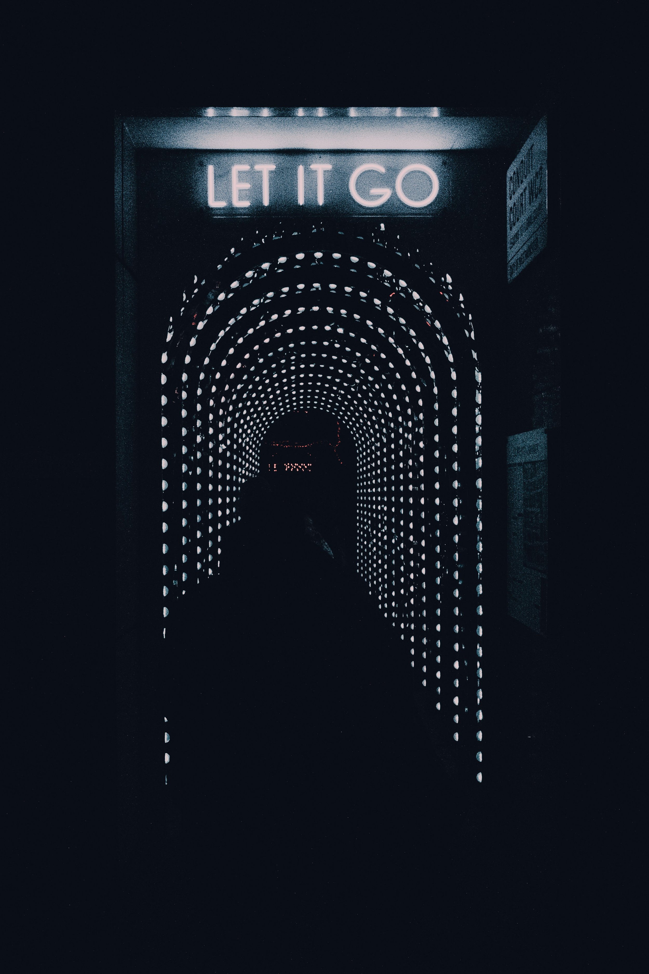 A dark, tunnel-like hallway lined with small circular lights forming an illuminated arch. At the entrance, a glowing neon sign reads “LET IT GO.” A shadowy figure stands at the opening, facing into the tunnel.