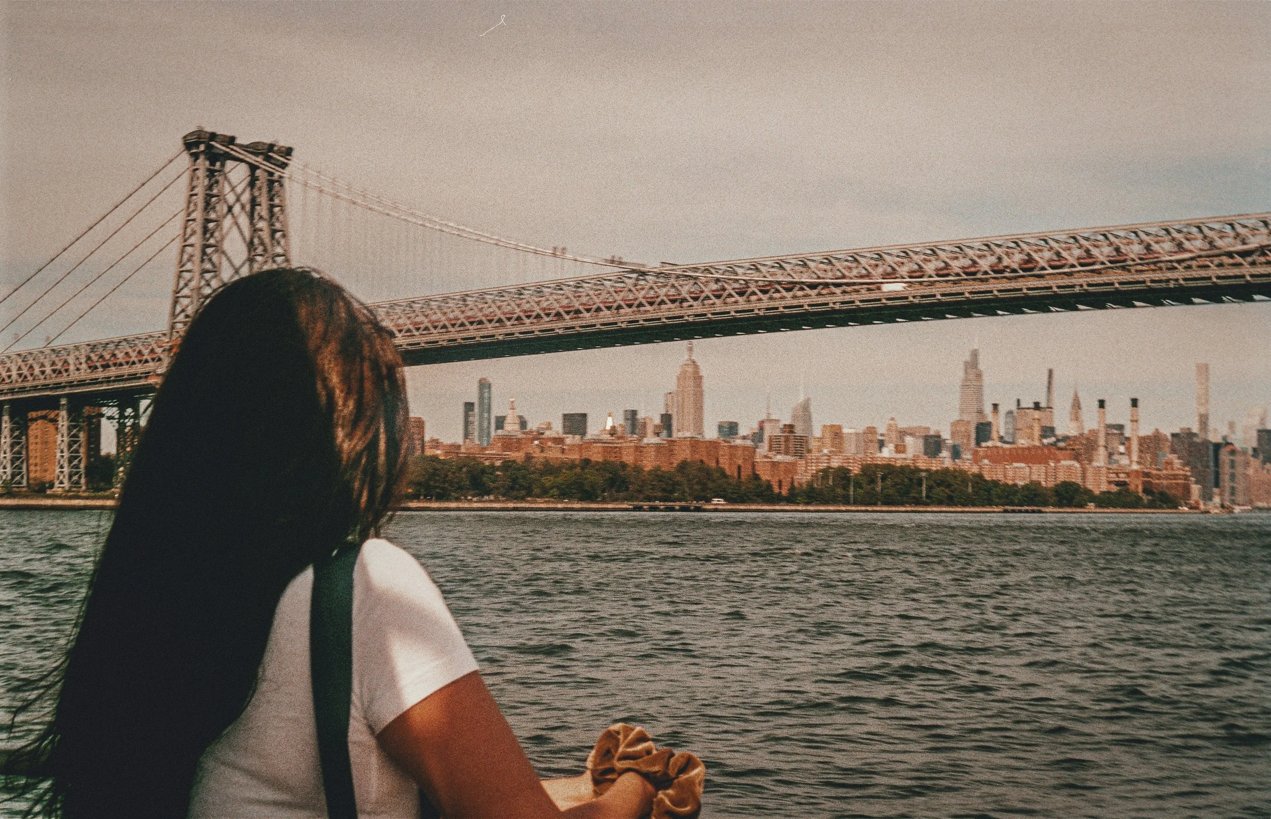 Person looking out at the NYC skyline and bridge, reflecting by the water.