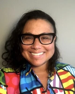 Smiling person with curly dark hair and black glasses wearing a colorful patterned shirt against a light background.