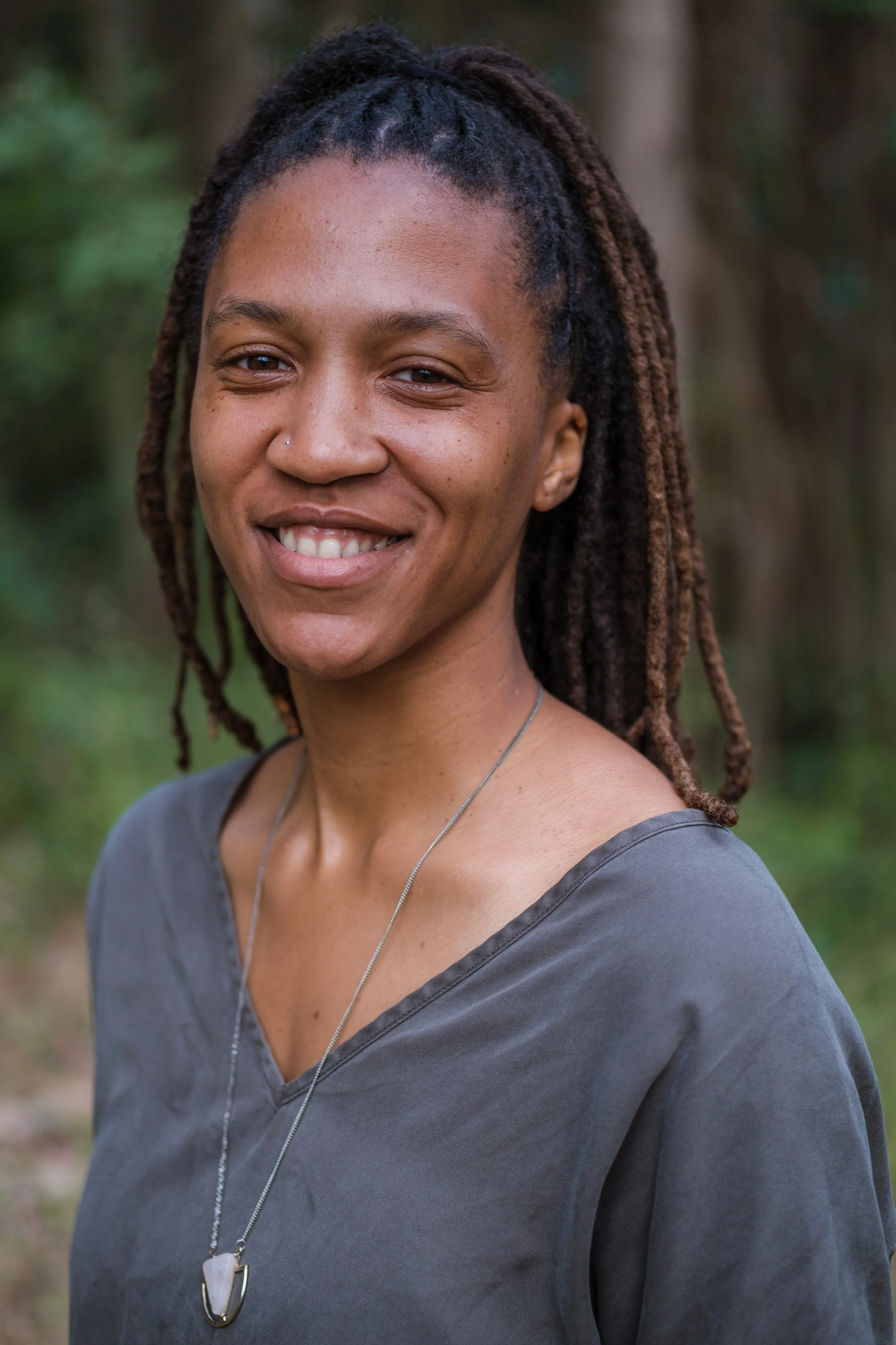 Smiling Black person with long locs pulled back, photographed outdoors against a softly blurred natural background.