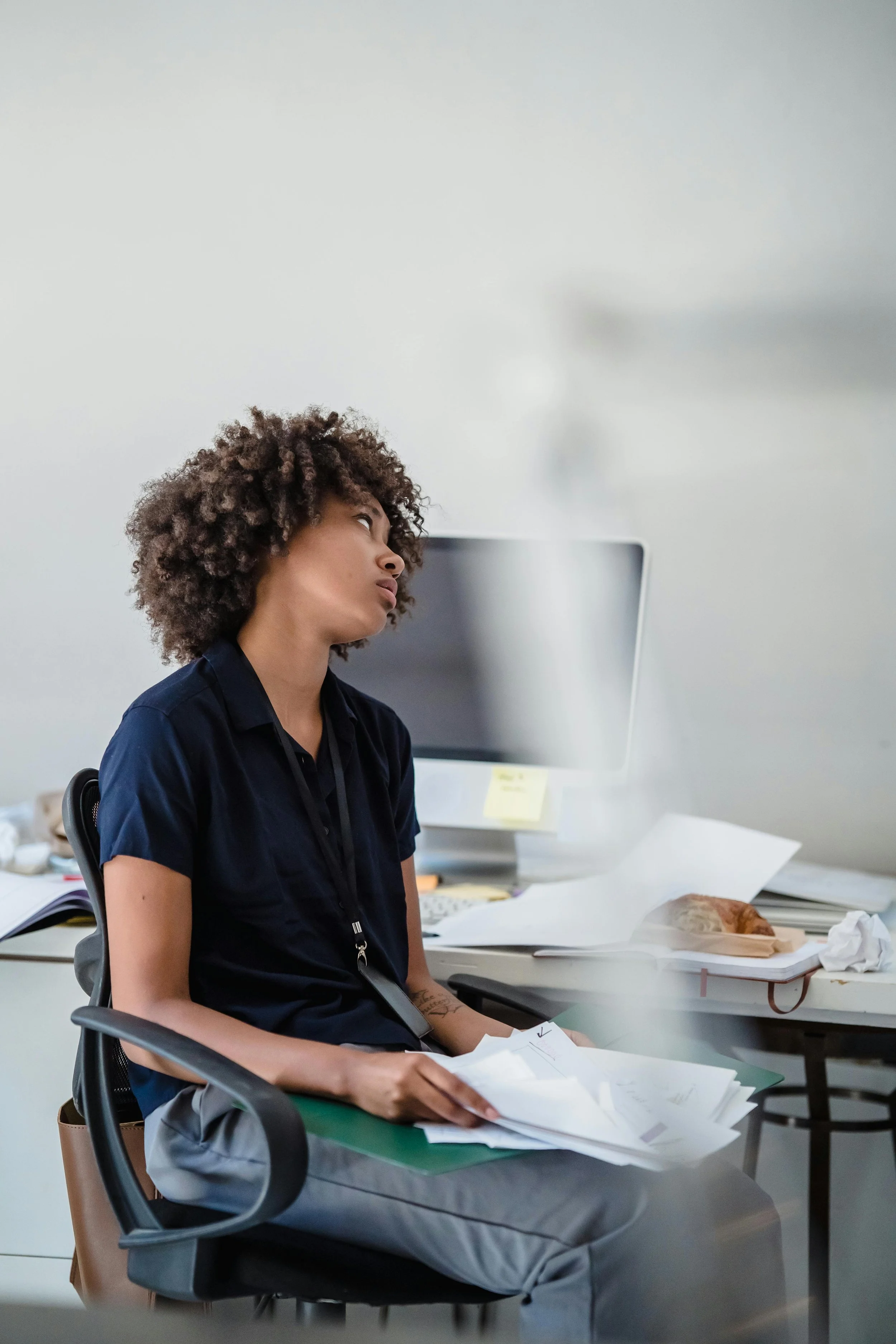 A tired-looking woman sits at her cluttered desk holding papers, gazing off in frustration or exhaustion.