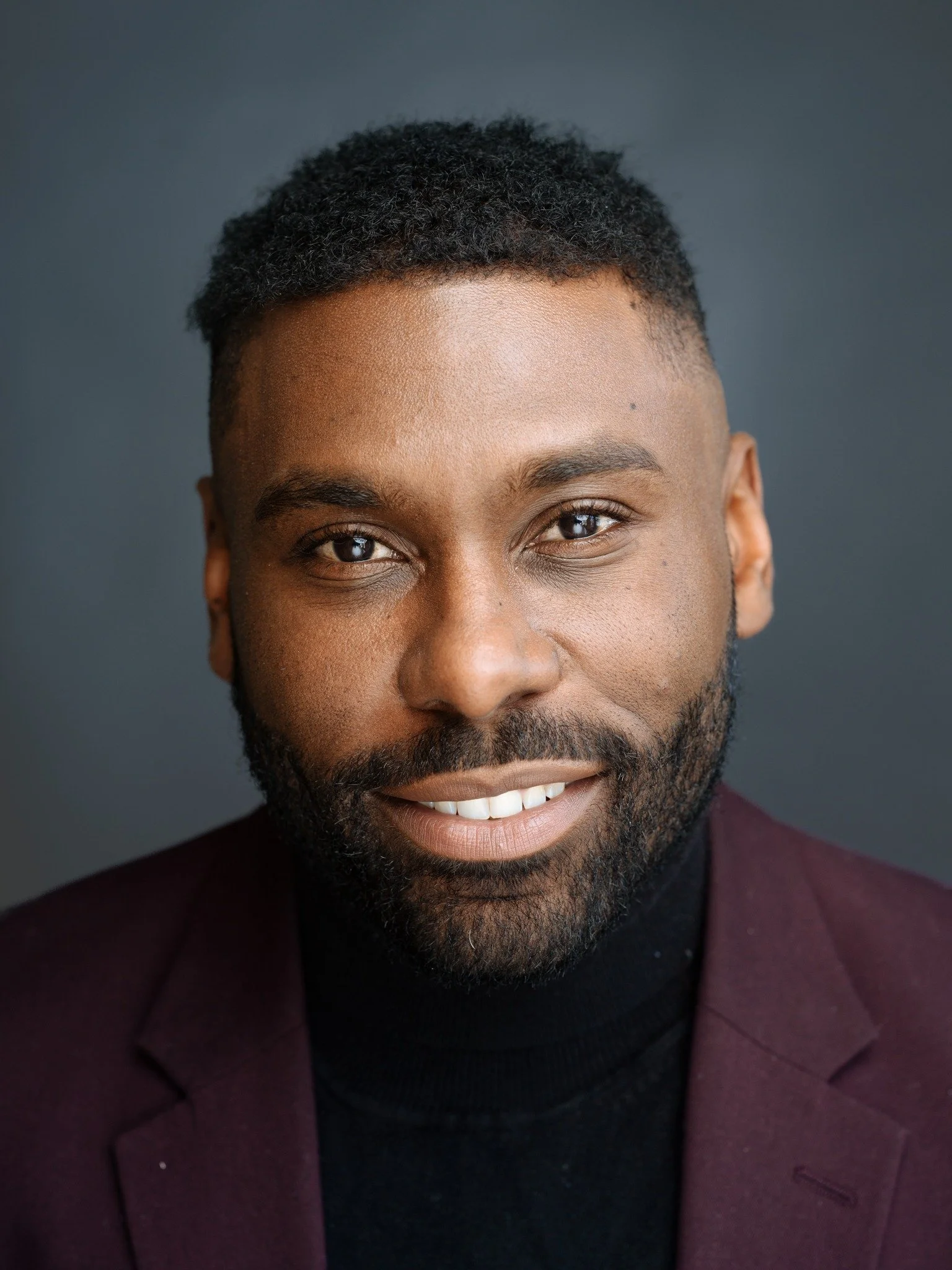 Color headshot of Marcus Stewart, smiling at the camera, with short hair and a full beard, wearing a burgundy blazer over a black turtleneck against a dark gray background