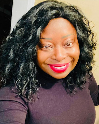 Photo of Nash Augustine, smiling at the camera, with long, voluminous curly black hair and bright red lipstick, wearing a plum turtleneck against a light cream wall.