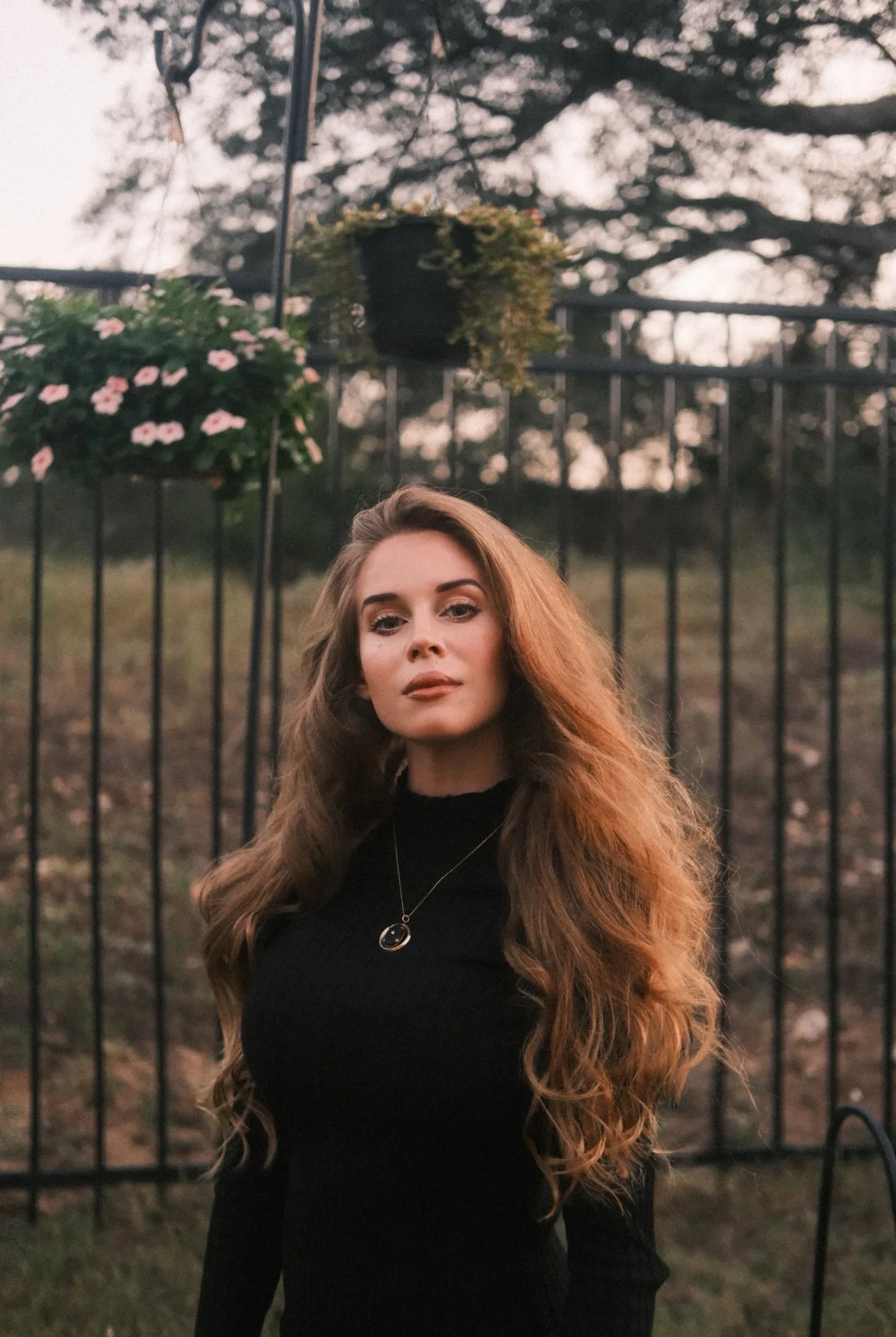 A person with long reddish blond hair wearing all black standing in front of an iron gate looking directly at the camera.