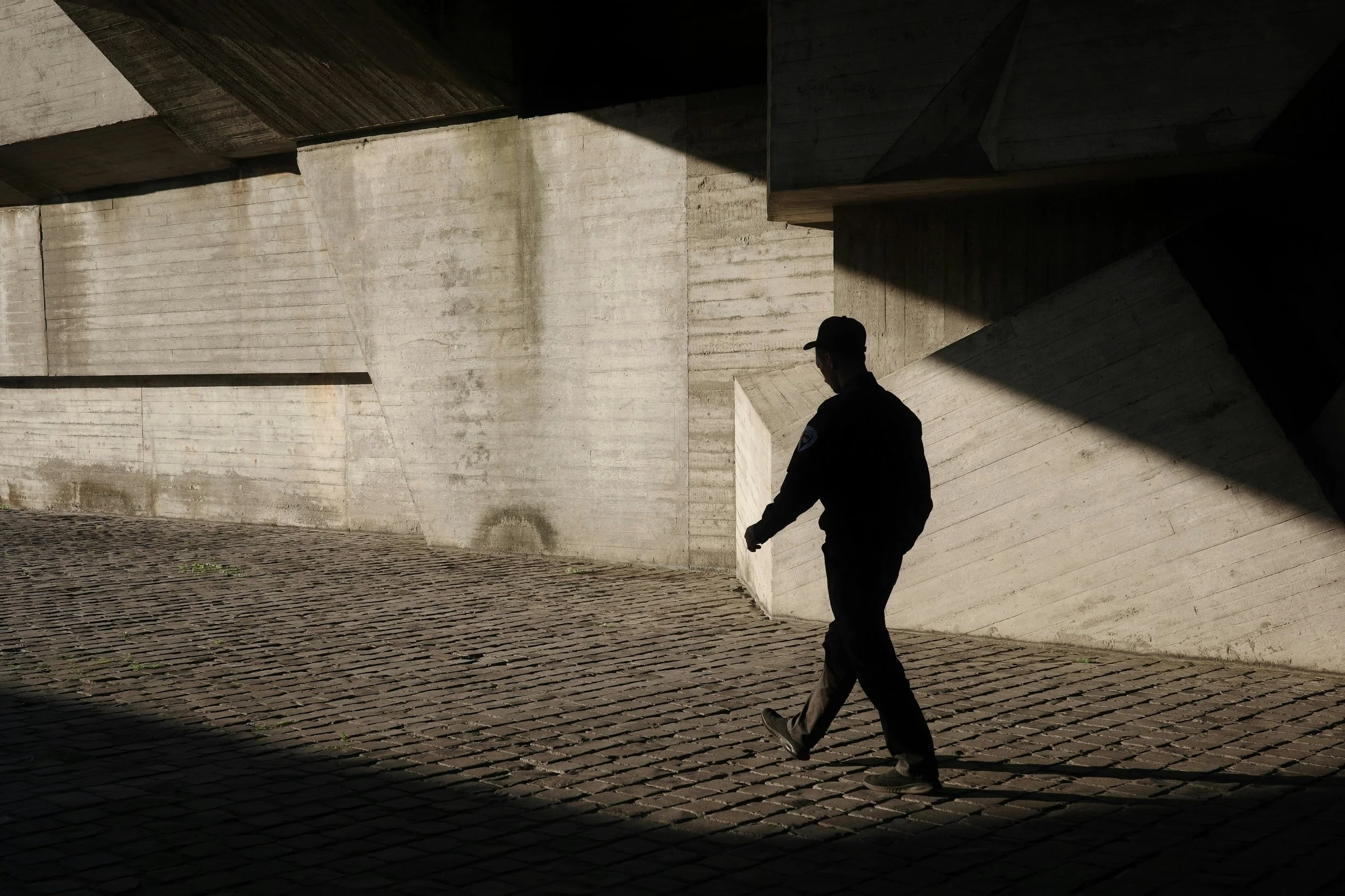 A person in dark clothing and a cap walks across a cobblestone path beside a large concrete wall. Strong sunlight creates sharp geometric shadows across the wall and ground, highlighting the figure’s silhouette in motion.