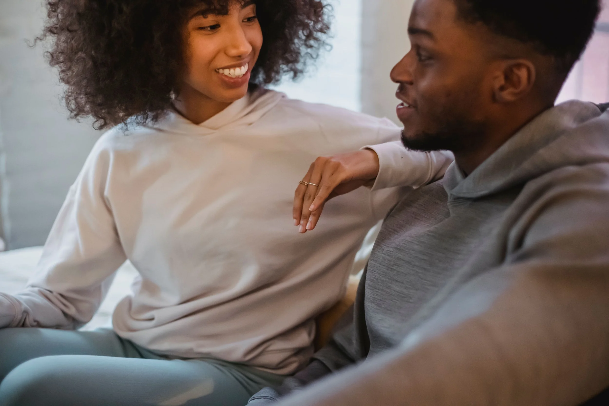 A black couple in cozy hoodies sits close on a couch, smiling and talking as she rests her arm on his shoulder.