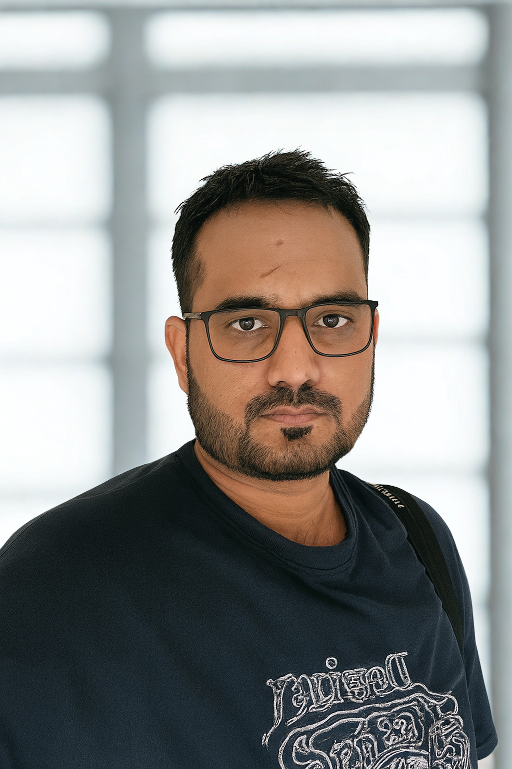 Man with short dark hair, beard, and glasses wearing a dark shirt with a graphic design, standing indoors near a textured wall and looking at the camera.