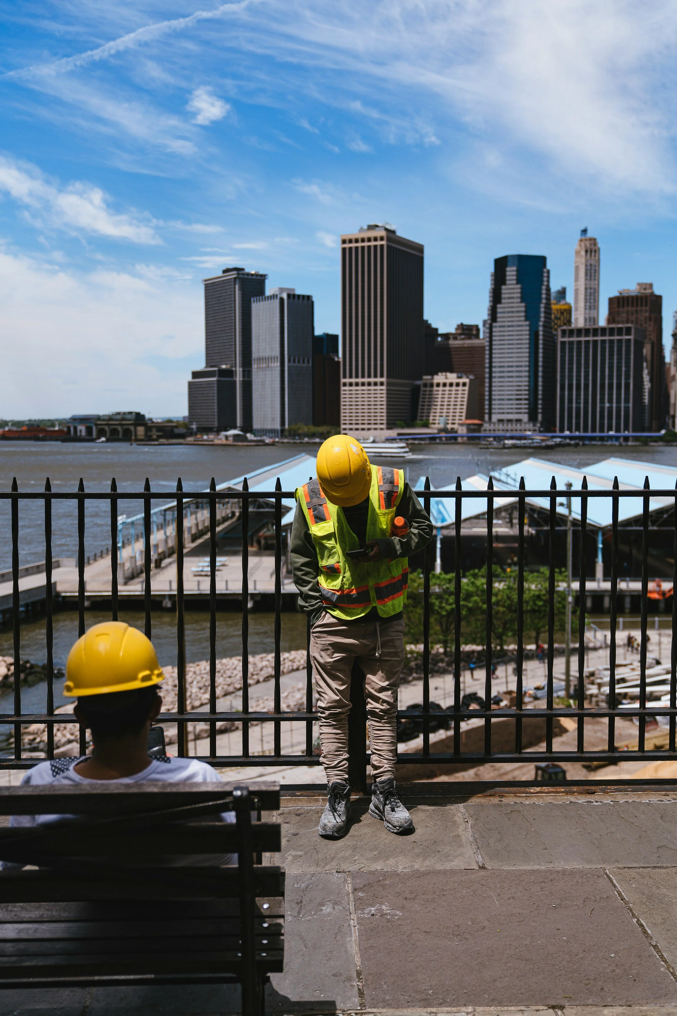 Photo of two construction workers, one is sitting on a bench looking at their phone while the other person is leaning against a gate with his hands in his pockets looking down. Behind him is the NY skyline.