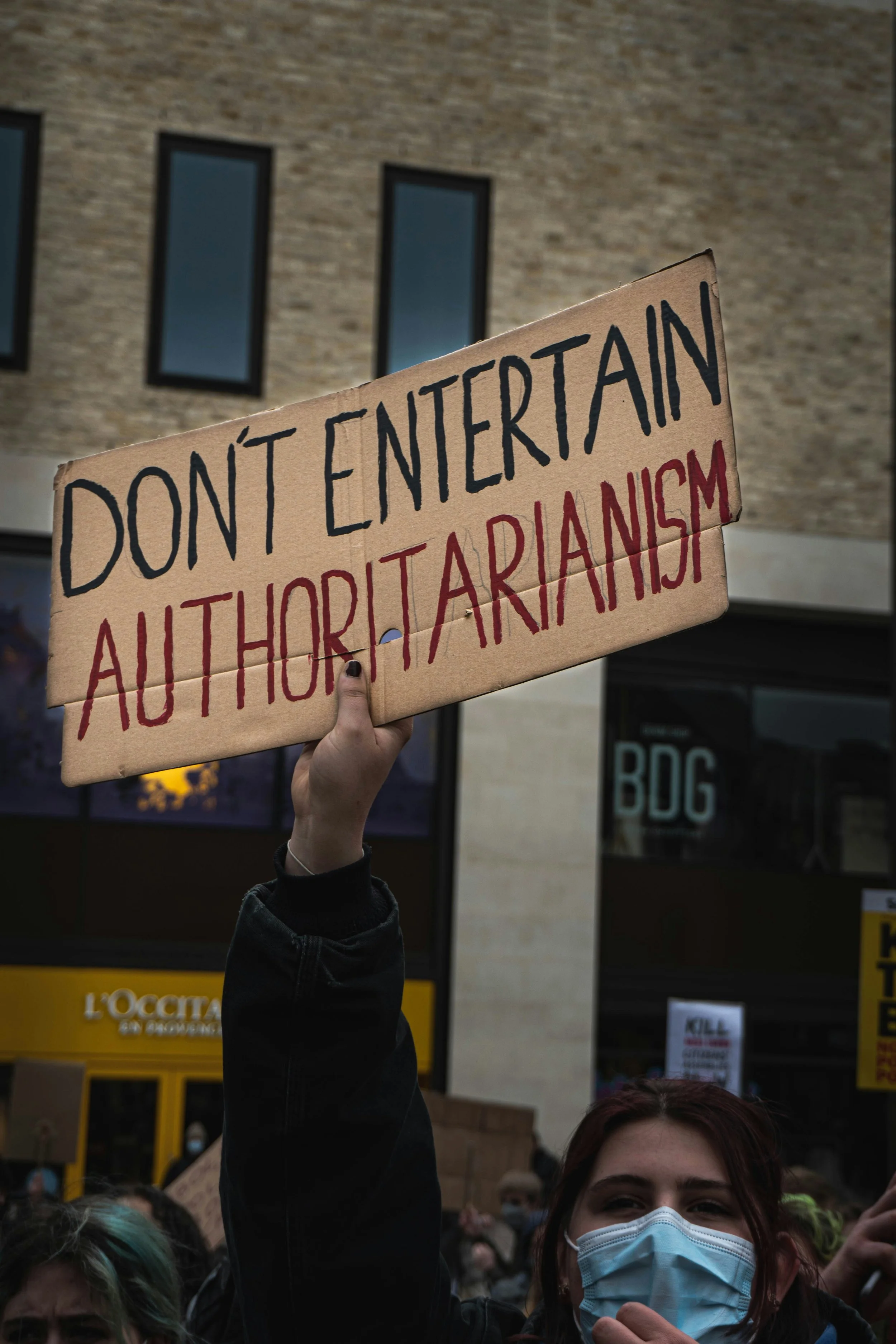 Photo of a woman wearing a face mask holding a sign that says "Don't Entertain Authoritarianism"