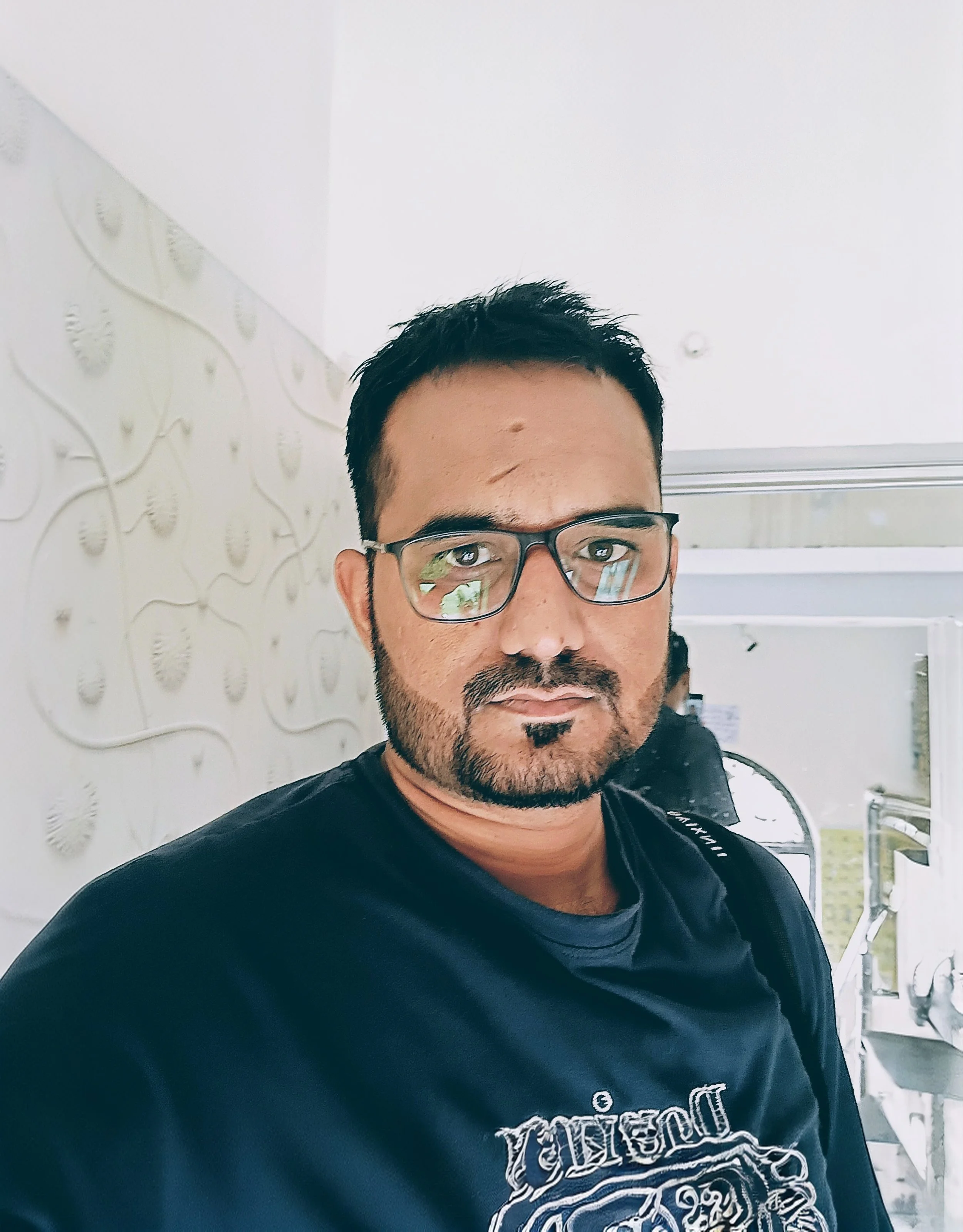 Man with short dark hair, beard, and glasses wearing a dark shirt with a graphic design, standing indoors near a textured wall and looking at the camera.