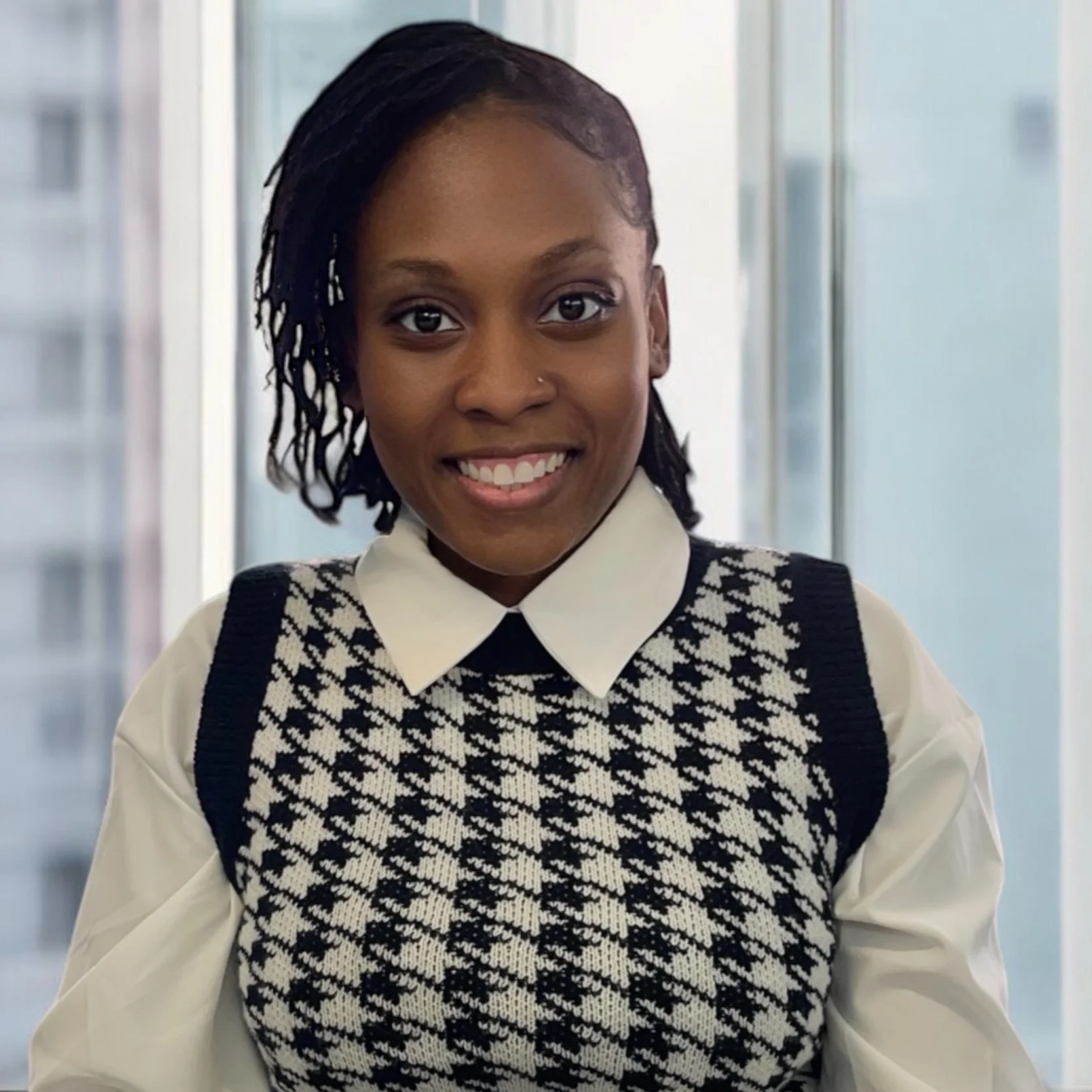 Smiling person in a white collared shirt and black-and-white patterned sweater vest, standing in a bright office.