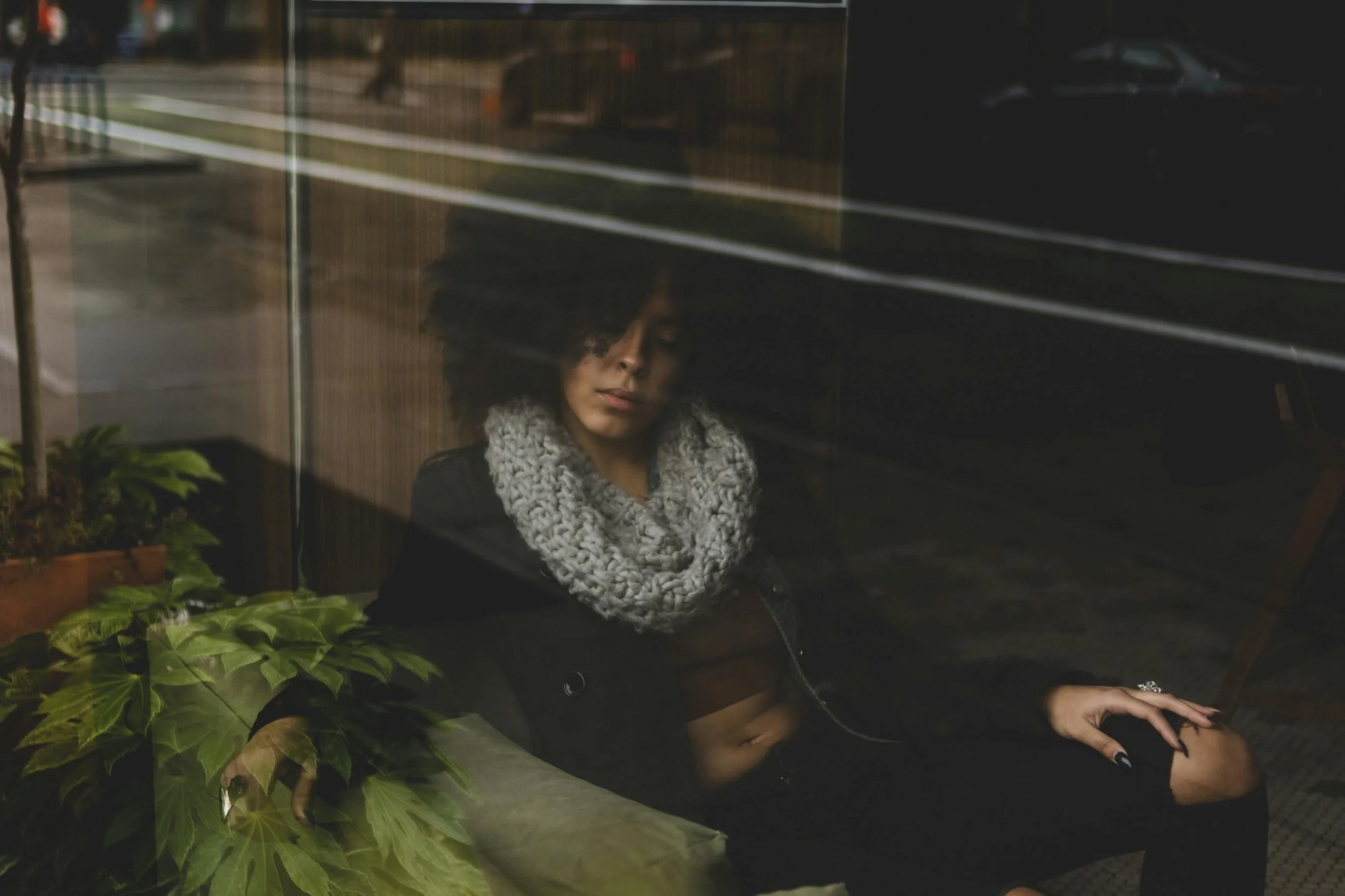 A photo of a person seated indoors behind a large window, partially obscured by dark glass reflections and streaks of city light. They wear a thick knit scarf and dark clothing, with one hand resting near green plants by the window.