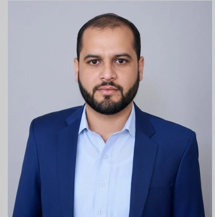 Man with short dark hair and beard wearing a blue suit jacket and light blue dress shirt, standing against a plain gray background with a neutral expression.