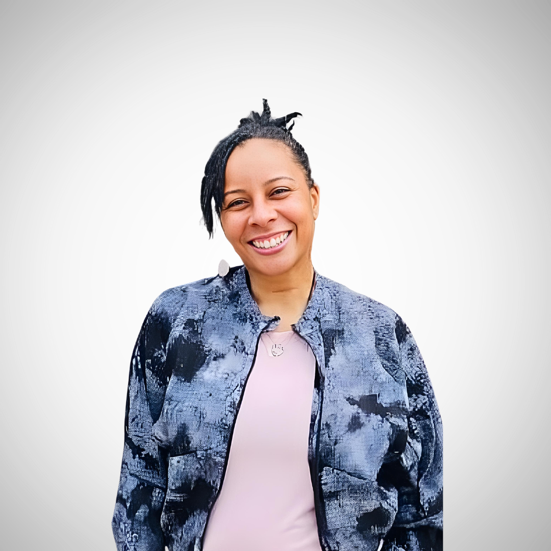 Smiling person with short locs wearing a light pink top and a blue-and-black patterned jacket, standing against a light gray background.