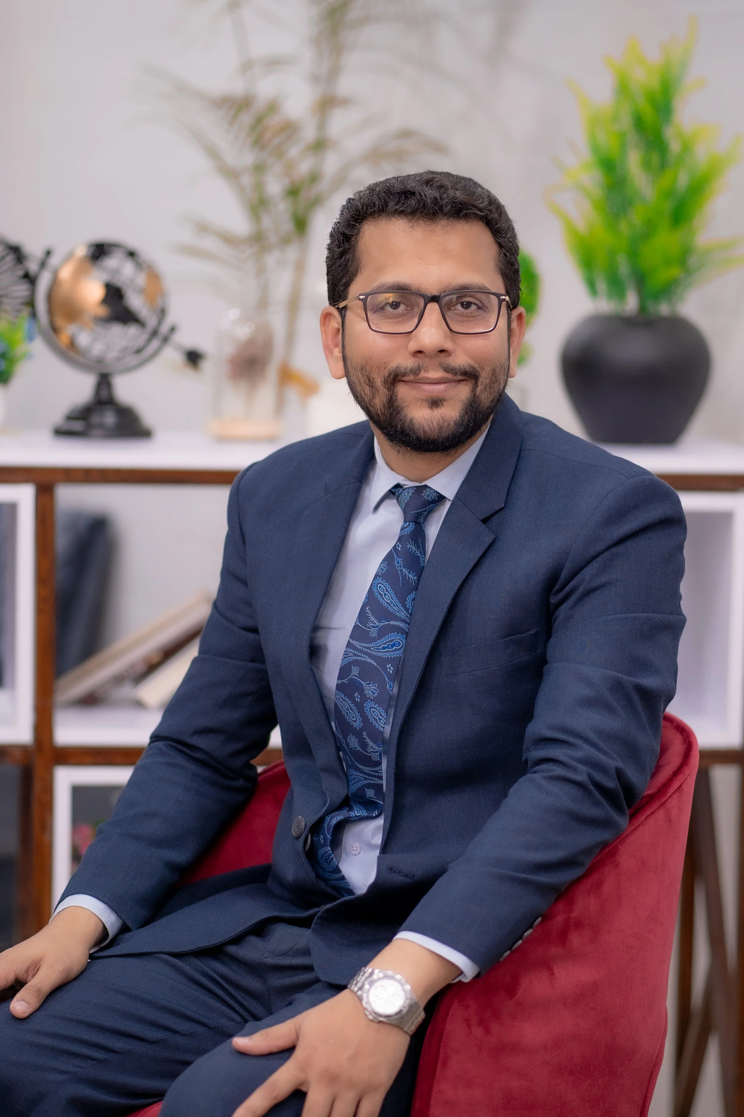 Man with short dark curly hair, beard, and glasses wearing a navy blue suit and patterned tie, sitting on a red chair and smiling slightly, with plants and a globe in the background.