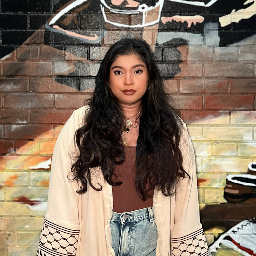 A person with long dark wavy hair stands facing the camera in front of a colorful painted brick mural, wearing a light cardigan over a brown top and high-waisted jeans.