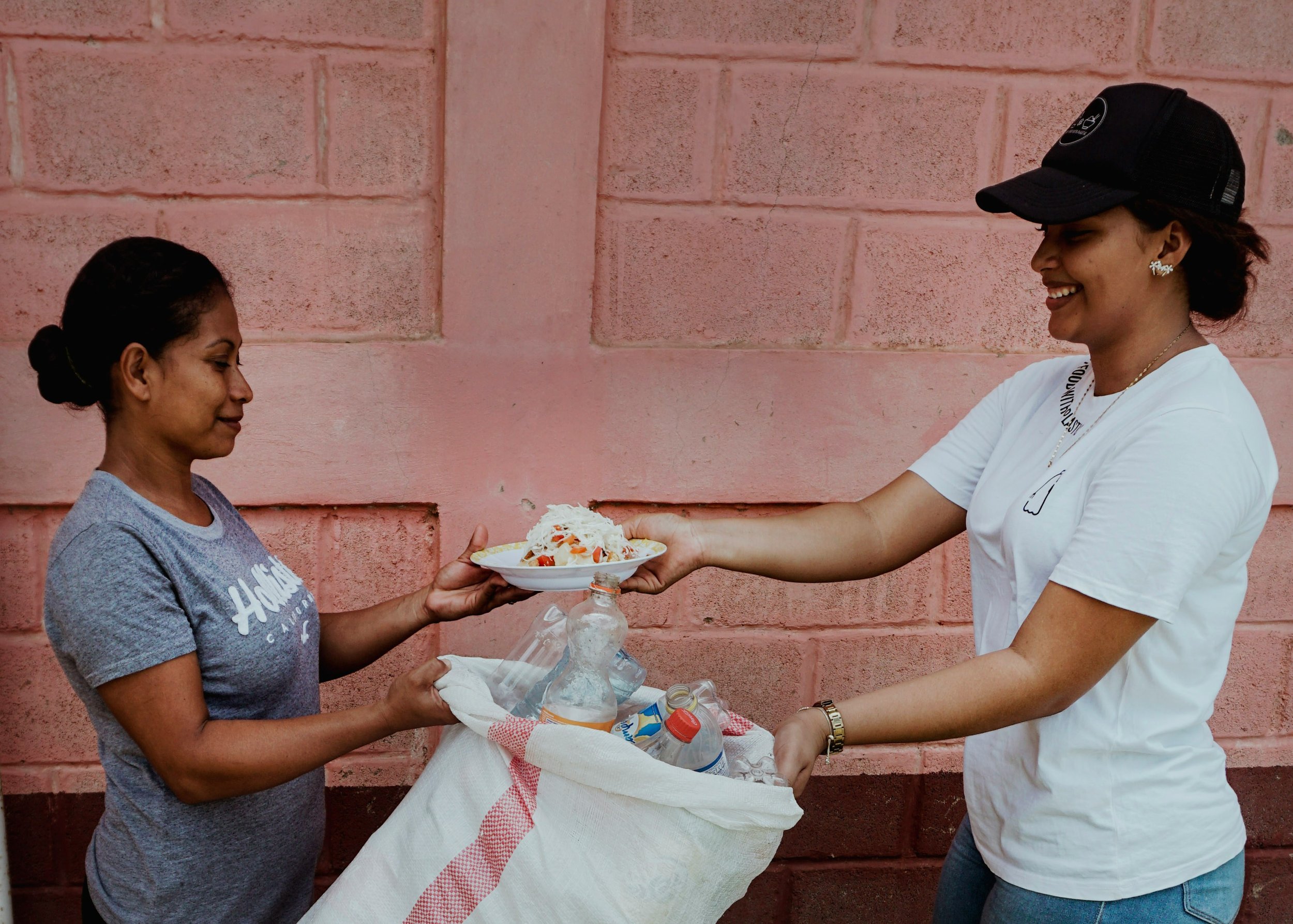 One woman handing a plate of food to another woman while carrying a bag of empty bottles, representing community care and sharing resources.