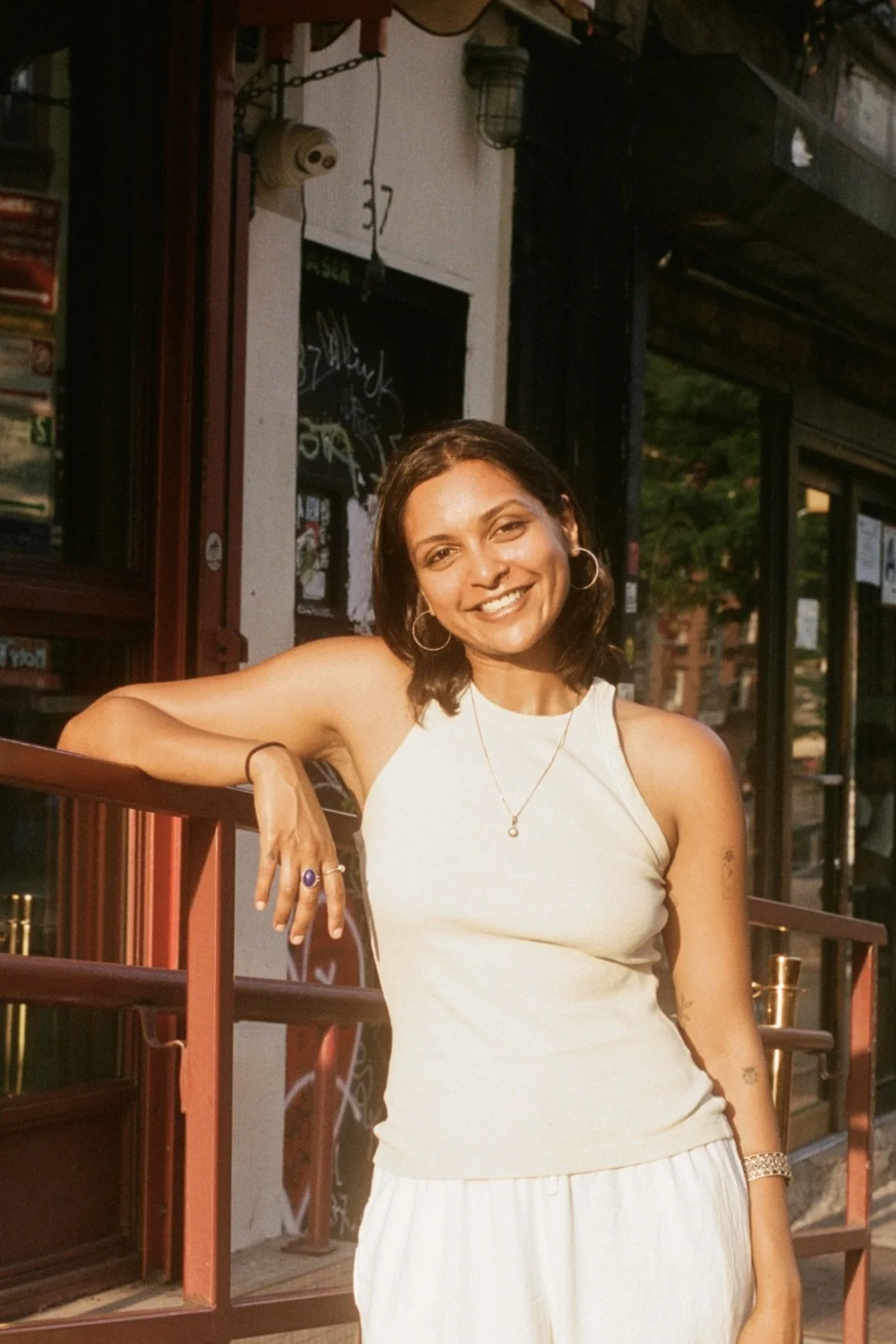 Photo of Riya Baphna, smiling and leaning on a railing outdoors in a warm sunlight, with shoulder-length wavy hair, gold hoop earrings and a pendant necklace, wearing a cream sleeveless top on a city street.