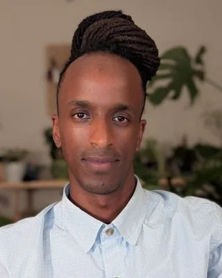 Person with locs in a bun. The person is wearing a baby blue button up and is sitting in front of plants.