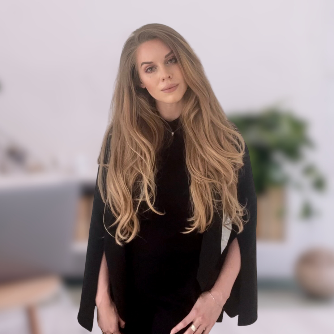 A person with long blonde hair wears a black top and black blazer, looking directly at the camera in front of a softly blurred office-style background with greenery.