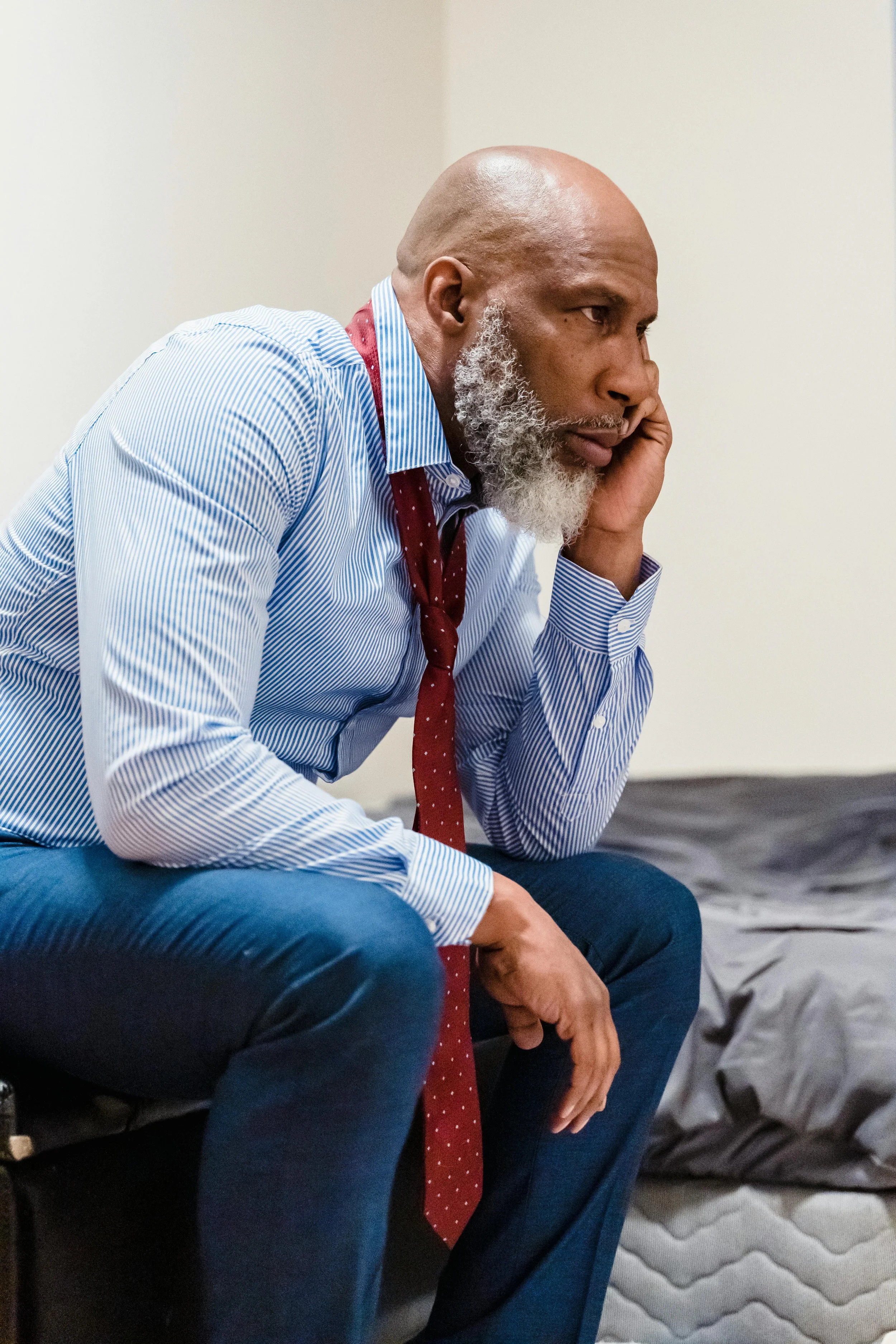 A man in a blue dress shirt and red tie sits on the edge of a bed, leaning forward with his hand on his face, appearing deep in thought or emotionally exhausted.