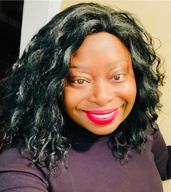 A smiling Black woman with curly black hair and bright red lipstick poses indoors wearing a dark turtleneck top.