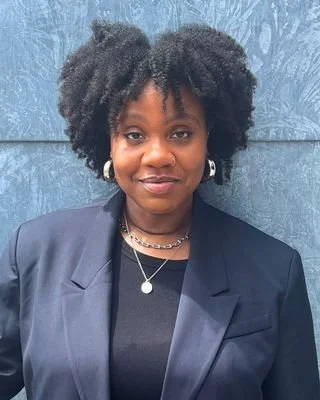 Headshot of Bianca Shaw, with voluminous natural curly hair and a soft, closed-mouth smile, wearing silver hoop earrings, layered silver necklaces, and a navy blazer over a black top against a textured blue-gray wall.