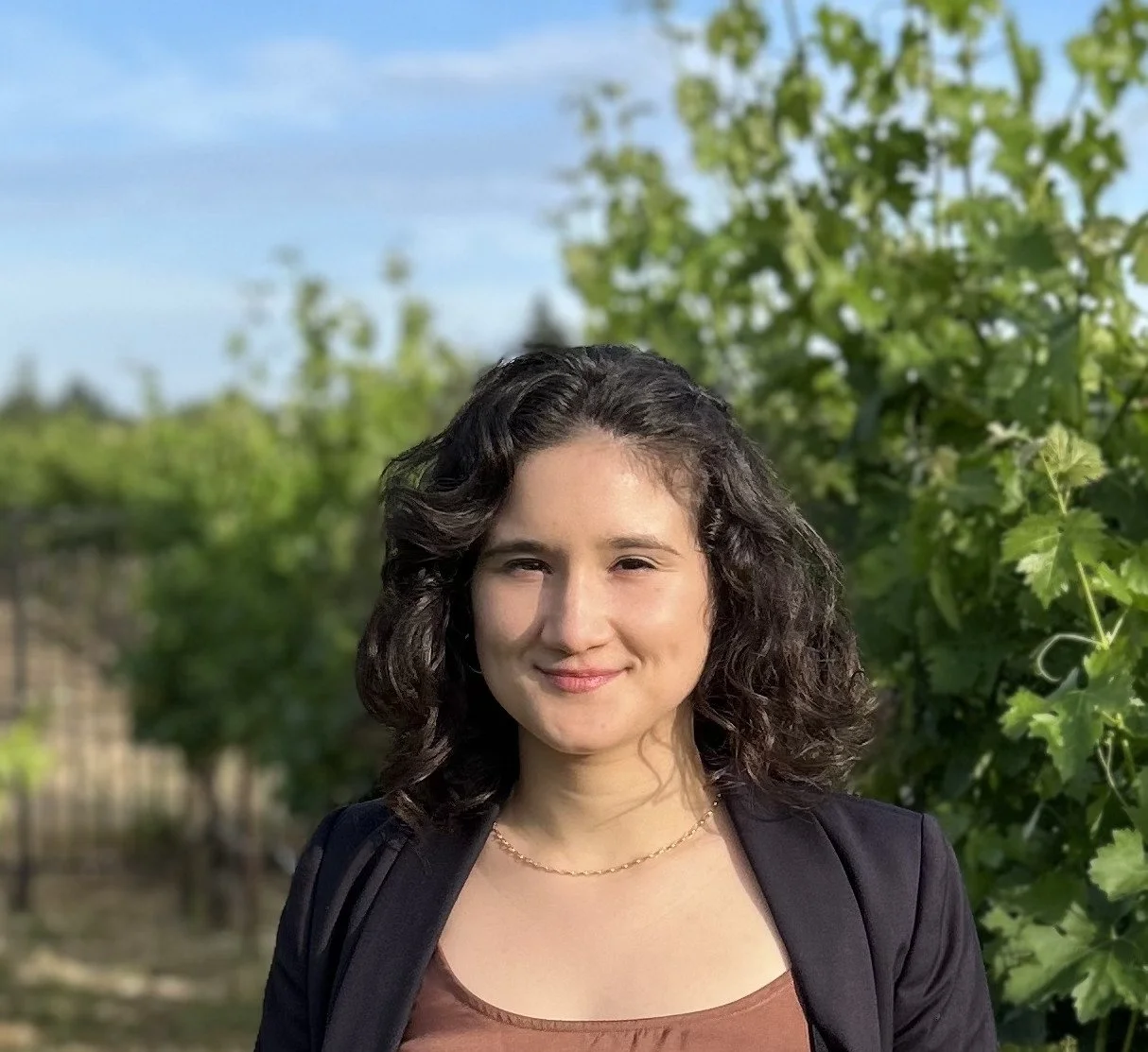 Headshot of Jess Miller-Suchet, smiling warmly outdoors, with shoulder-length dark curly hair, wearing a black blazer over a rust-colored top, with green leafy vines and a soft blue sky behind her.