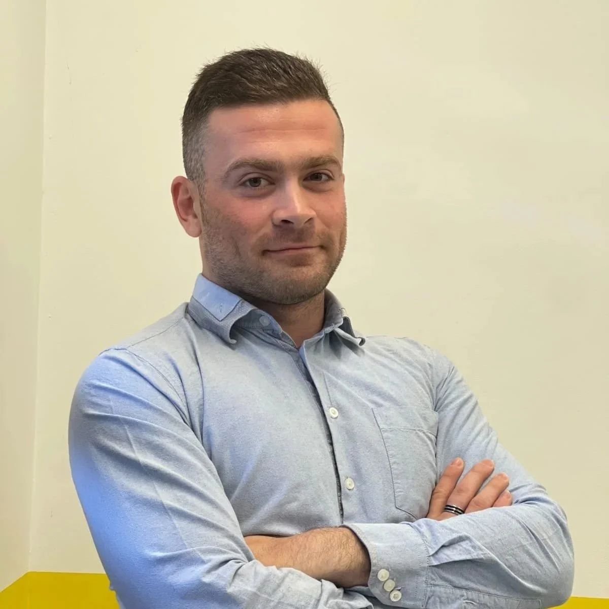 A man with short dark hair and a trimmed beard stands against a two-tone white and yellow wall. He is wearing a light blue button-up shirt and has his arms crossed, looking confidently at the camera.