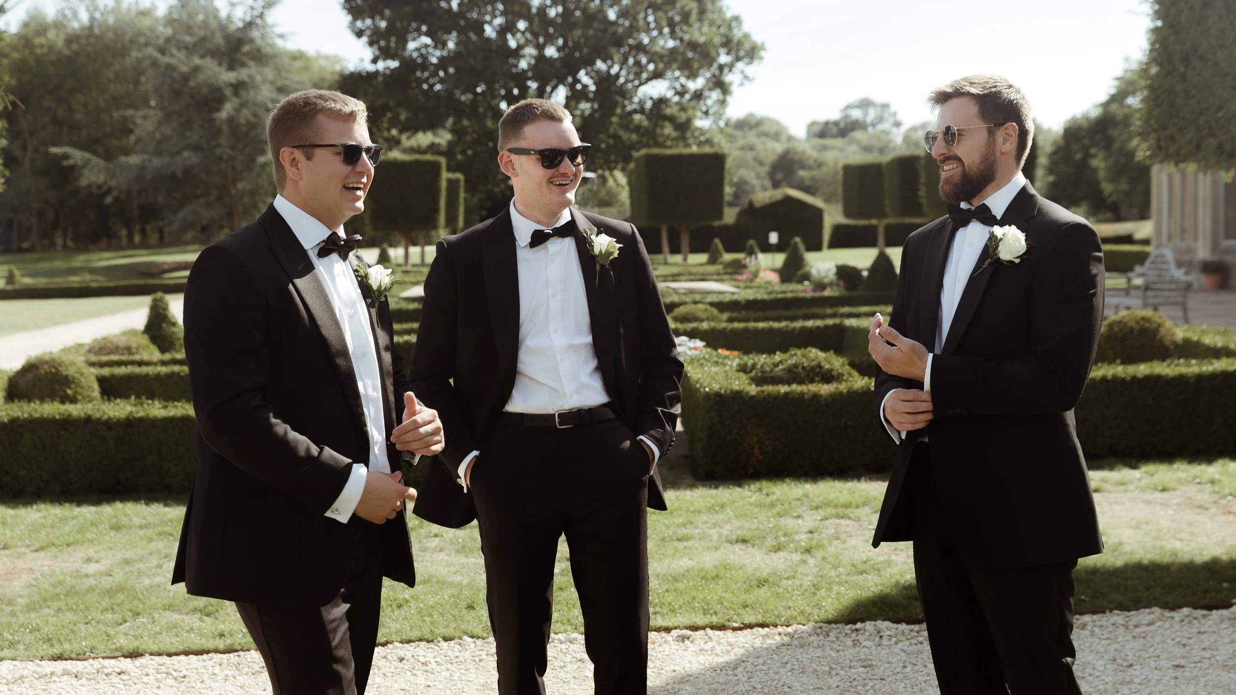 Three men in formal black tuxedos with white shirts and black bow ties, wearing sunglasses and white boutonnieres, are talking and smiling outdoors in a garden area with neatly trimmed bushes and trees.