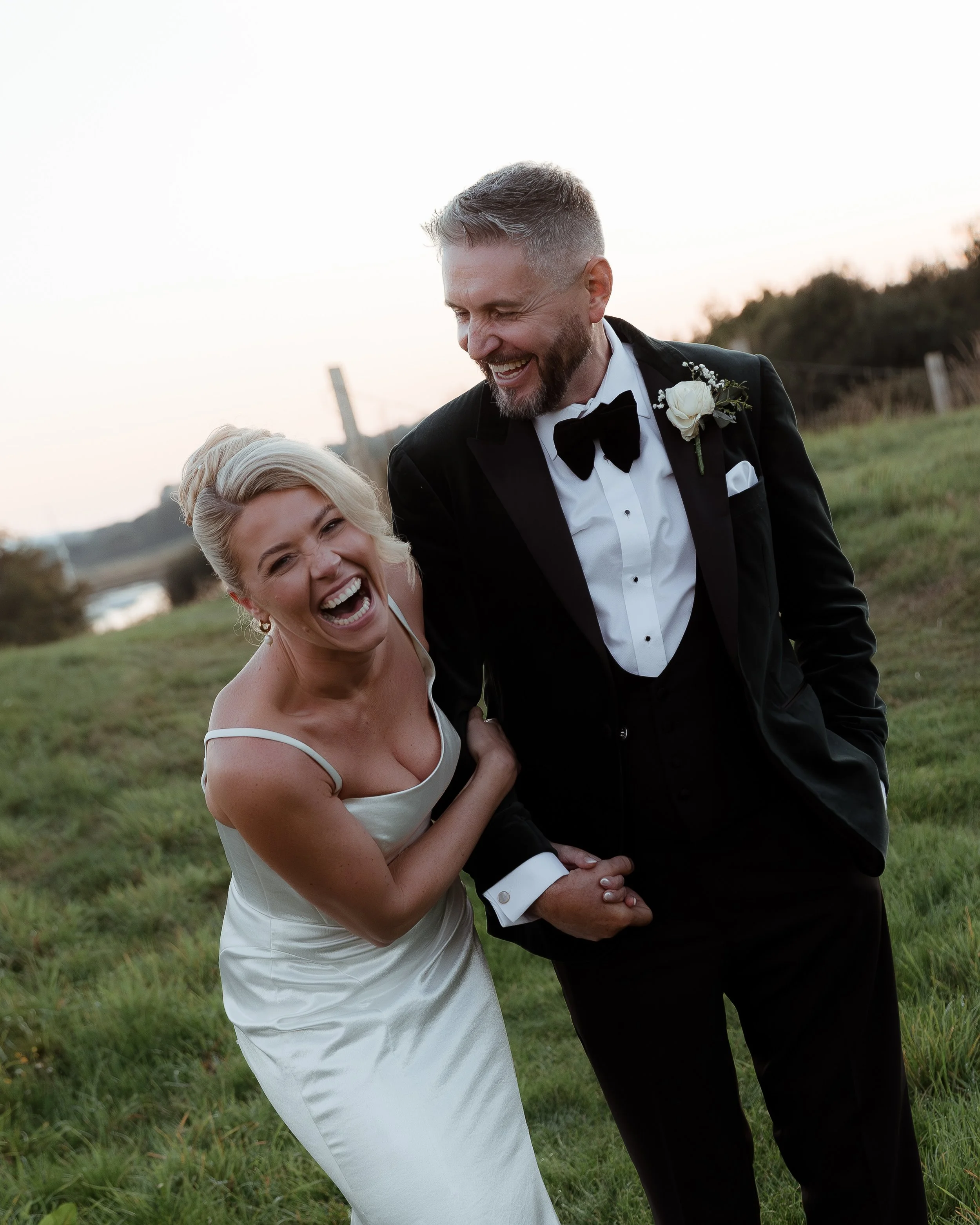 A bride and groom in wedding attire laughing and holding hands outdoors during sunset.