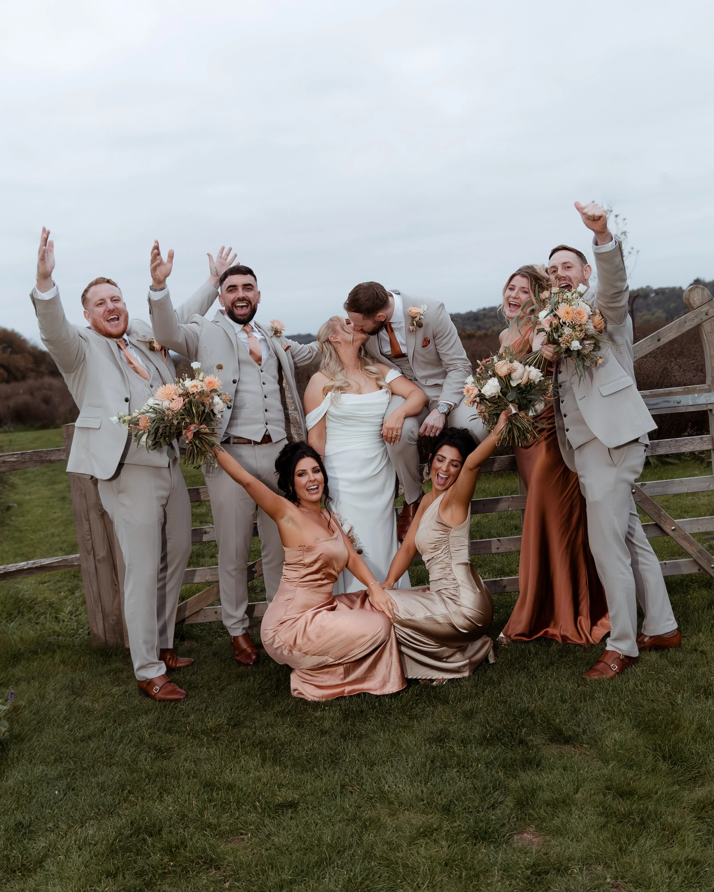 Group of wedding party members celebrating outdoors, with the bride and groom in the center, surrounded by bridesmaids and groomsmen holding bouquets, all smiling and raising their arms.
