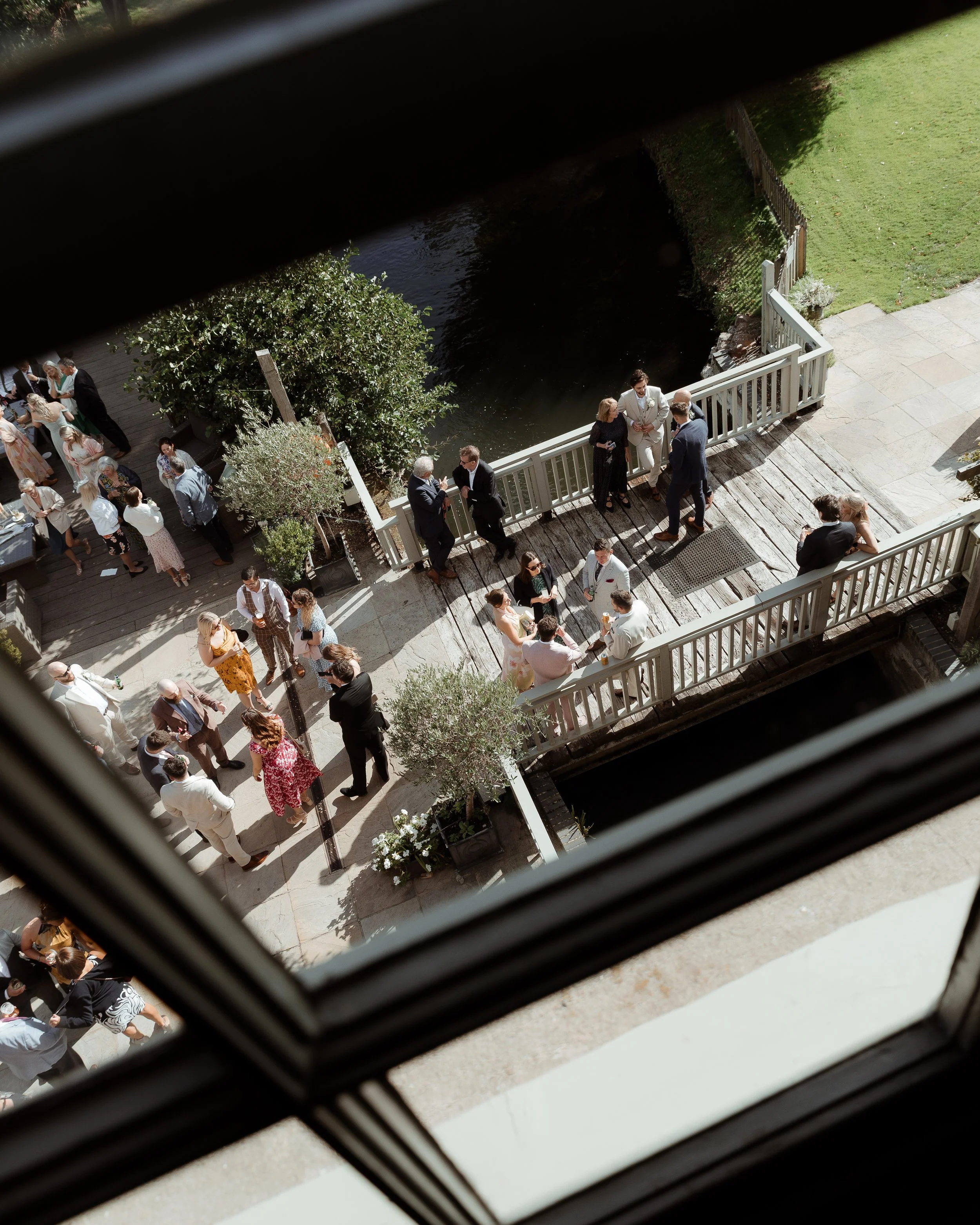 Guests at a wedding reception on a wooden patio overlooking a pond, with some guests talking near the railing and others gathered under trees and around tables.
