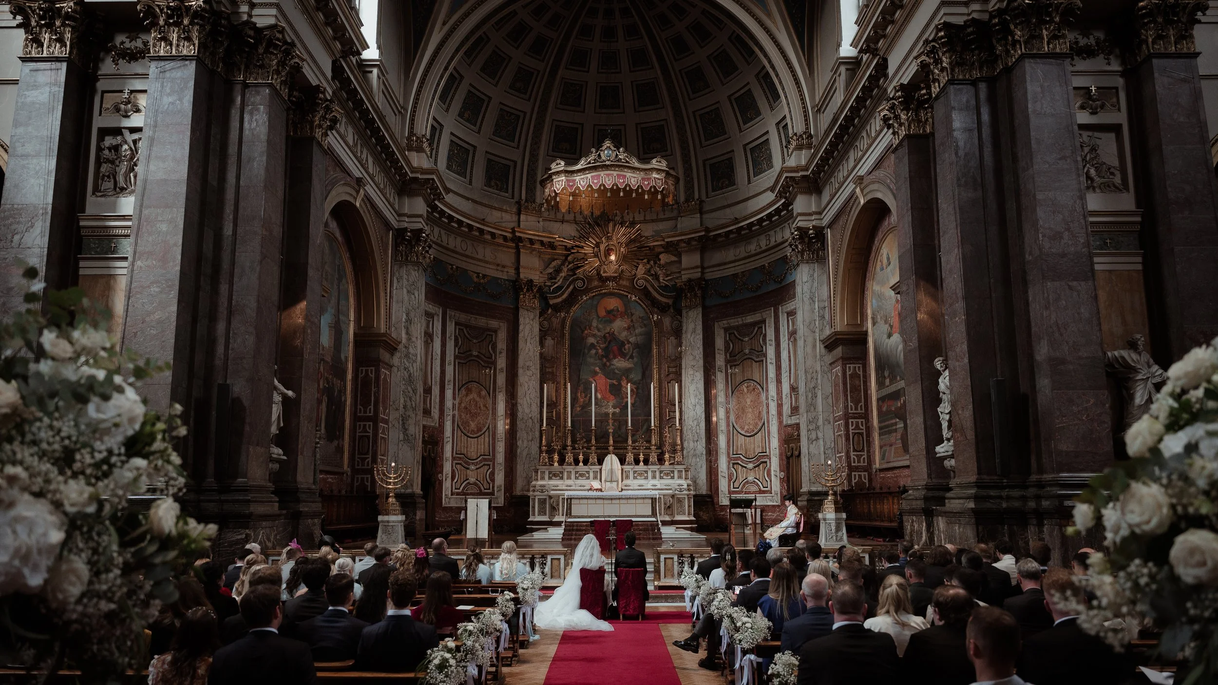 A wedding ceremony inside a grand, ornate church with numerous guests seated, a bride and groom kneeling at the altar, surrounded by flowers and religious artifacts.