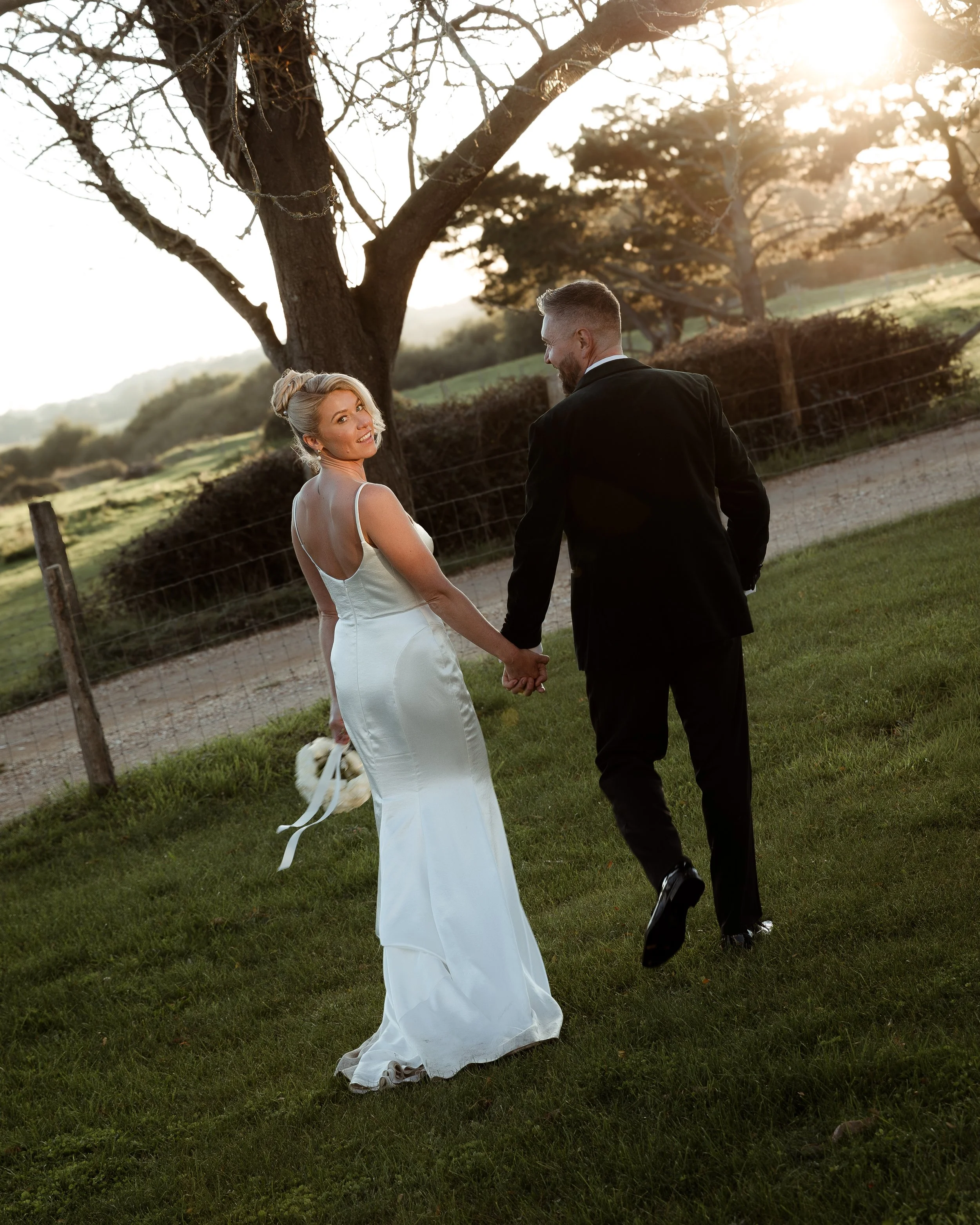 A bride holding a bouquet and a groom holding her hand walking outdoors during sunset in a grassy area with trees.