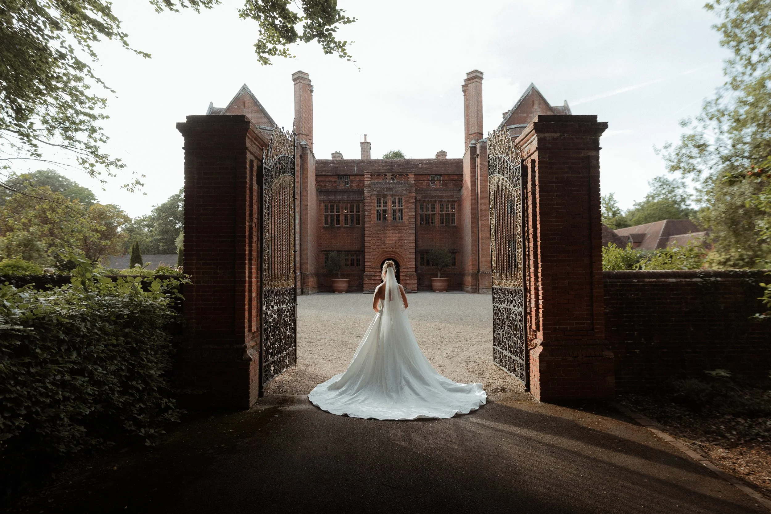 A bride in a white wedding gown with a long train standing at an open iron gate in front of a large, old brick estate or mansion surrounded by trees.