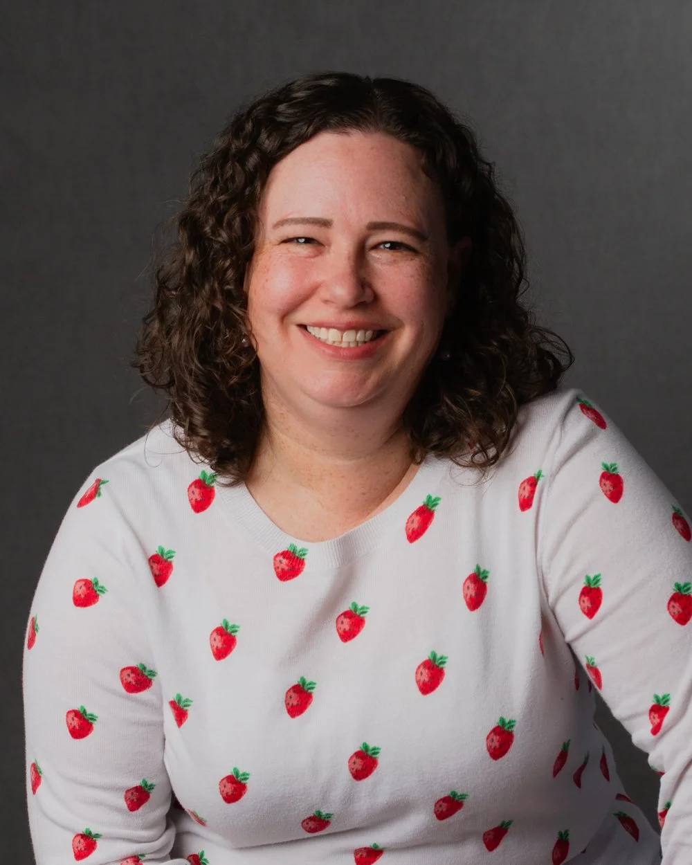 Photo of Lara Zelman smiling at the camera, wearing a white sweater with strawberry print.