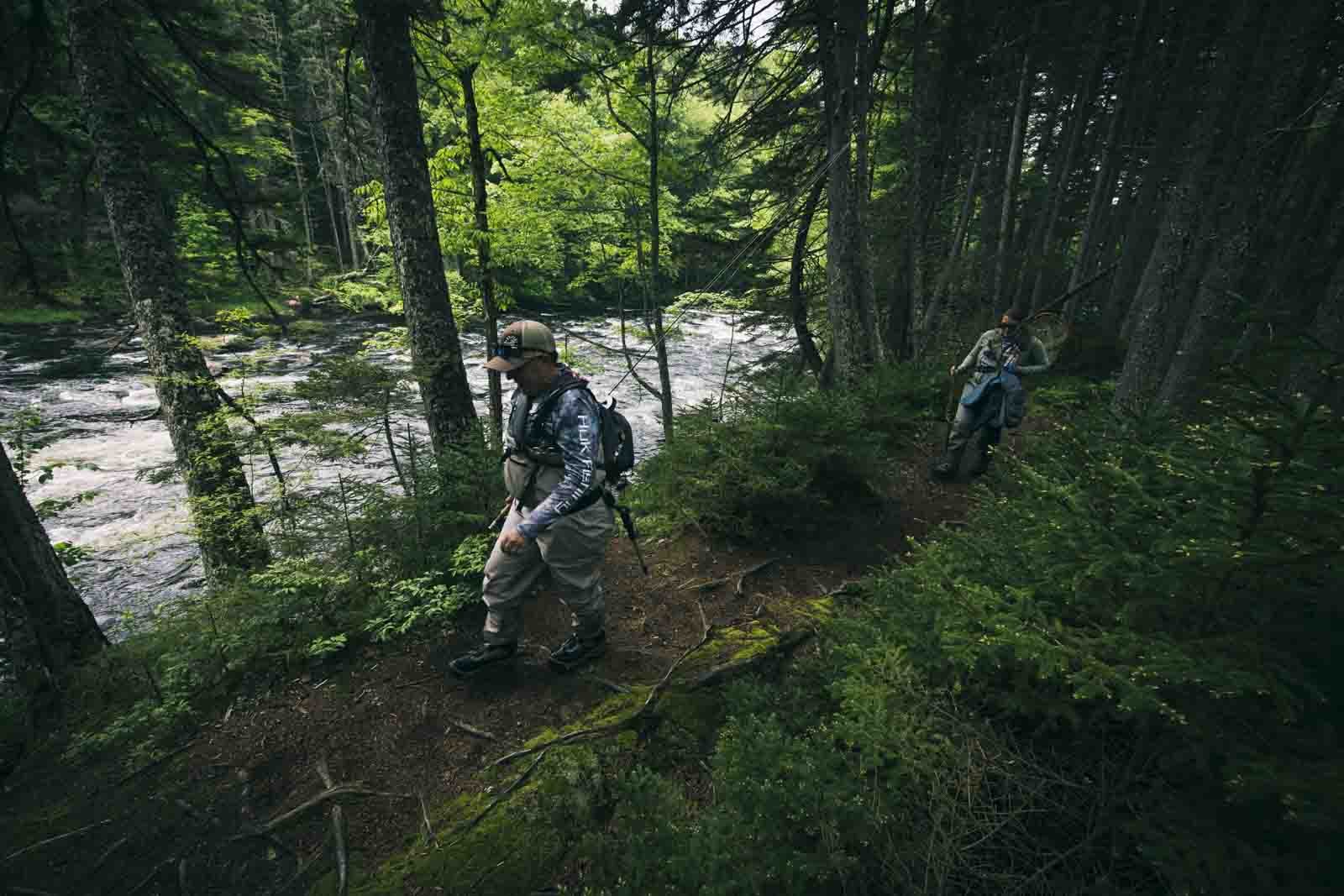  Bev (she/her) and  Megan  (she/her) walk alongside a river in western Maine, following a trail through conifers and undergrowth. Photo:  Joe Klementovich  