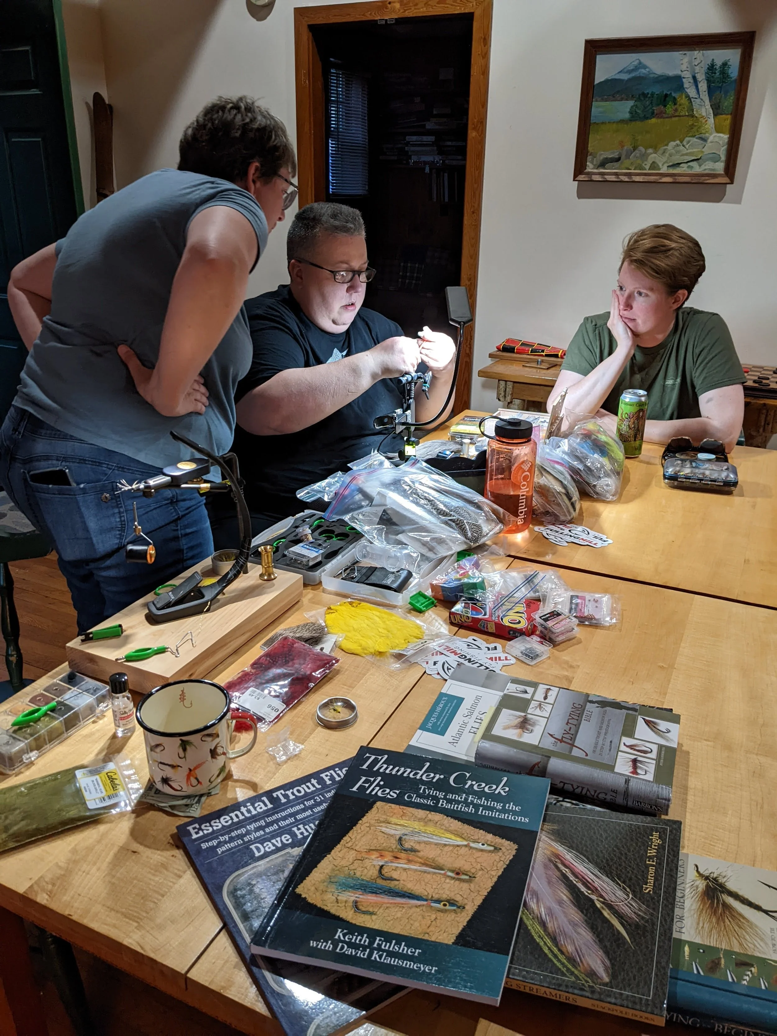 fly tiers gather inside at a large table with tying materials scattered amongst books on bugs and flies. Another camper watches as others tie at the vise.